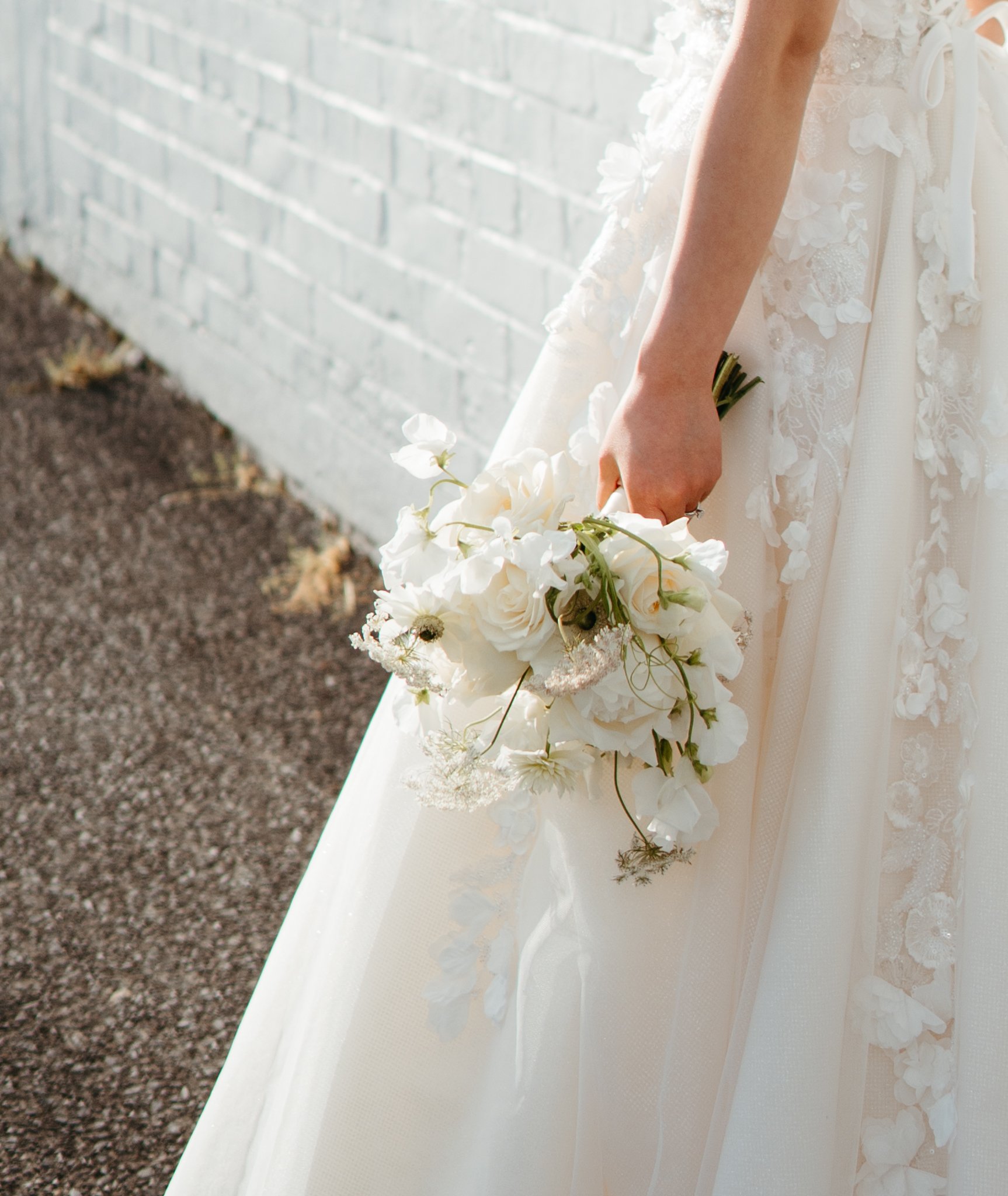 A bride in a white wedding dress holding a bouquet of white flowers, standing outdoors near a white brick wall.