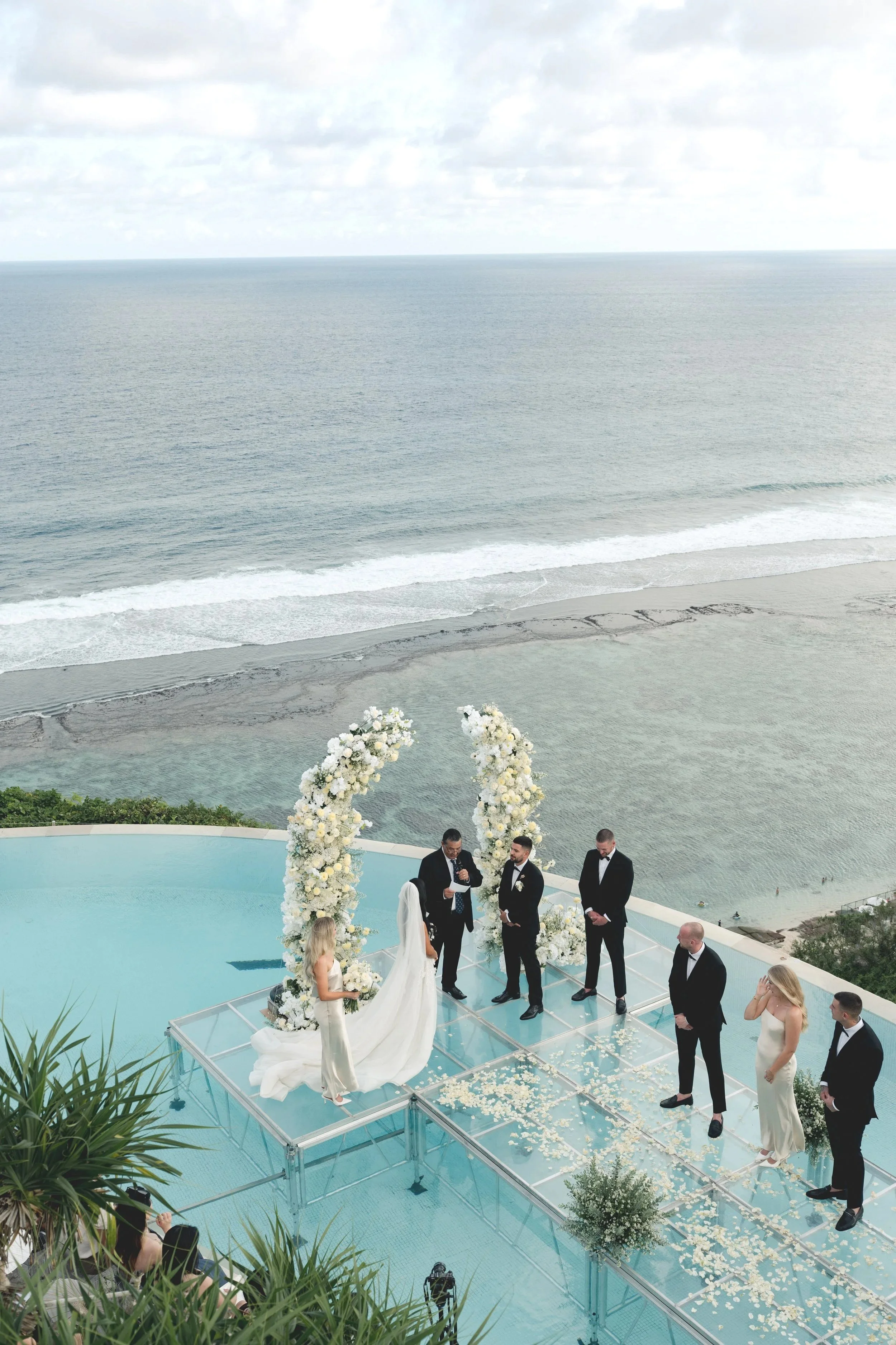 A wedding ceremony taking place on a glass platform near the beach with ocean views, decorated with white floral arches and scattered flower petals.