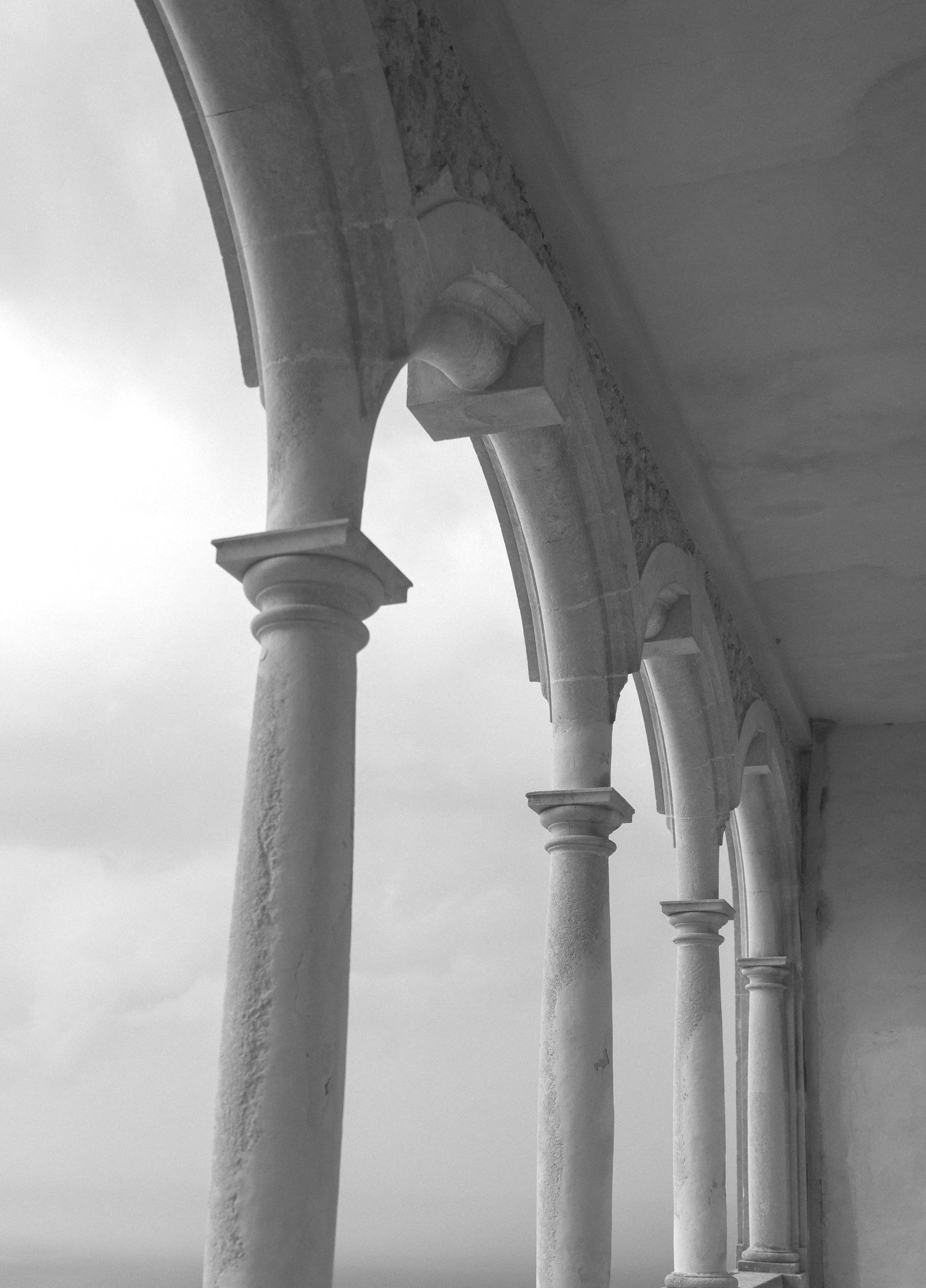 Black and white photograph of a stone balcony with decorative balusters and a curved railing, viewed from below against a cloudy sky.
