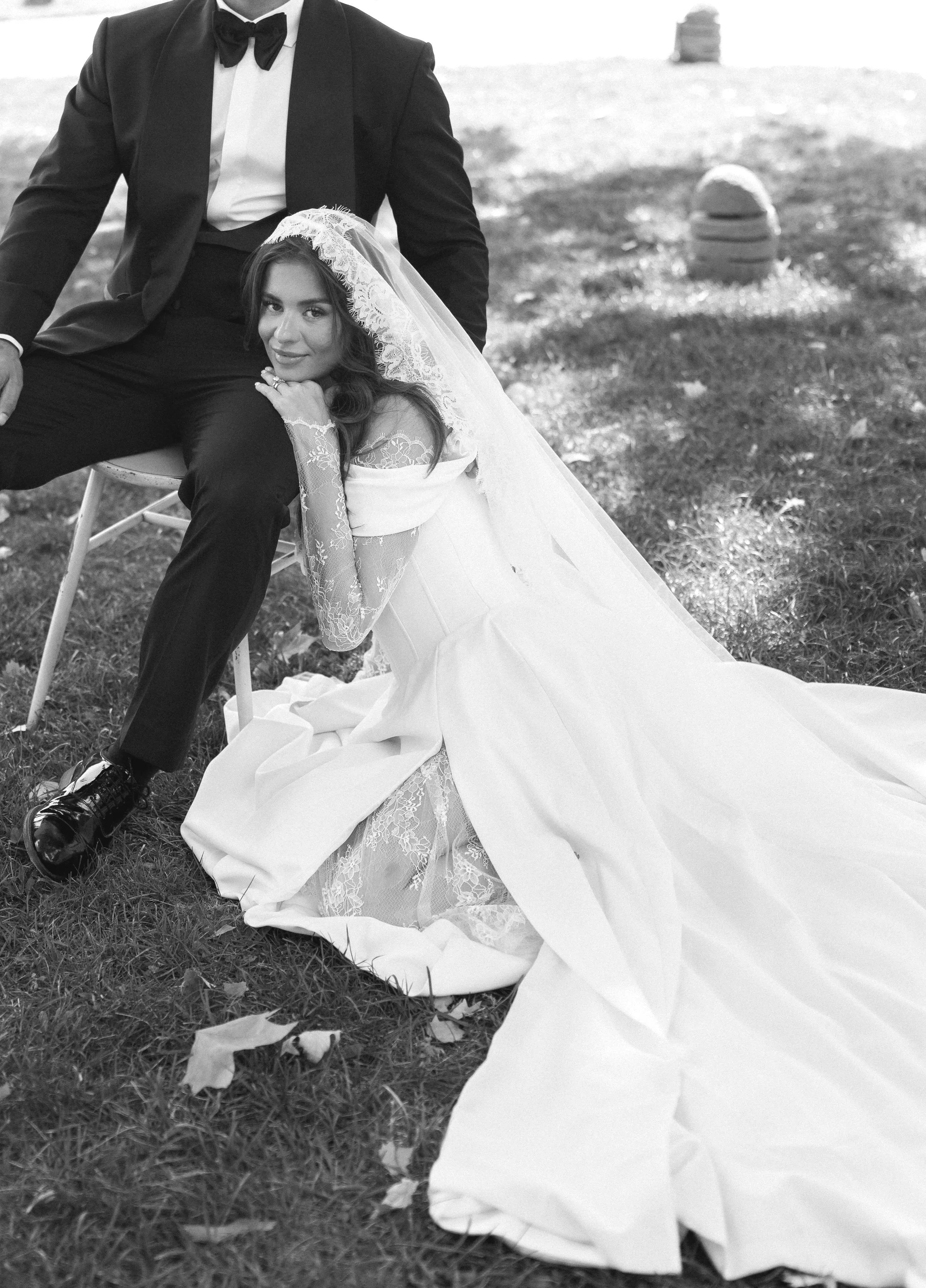 A woman in a wedding dress lying on the grass, resting her head on a man's lap, with a man dressed in a tuxedo sitting next to her. The scene is outdoors on a grassy area with a barrel in the background.