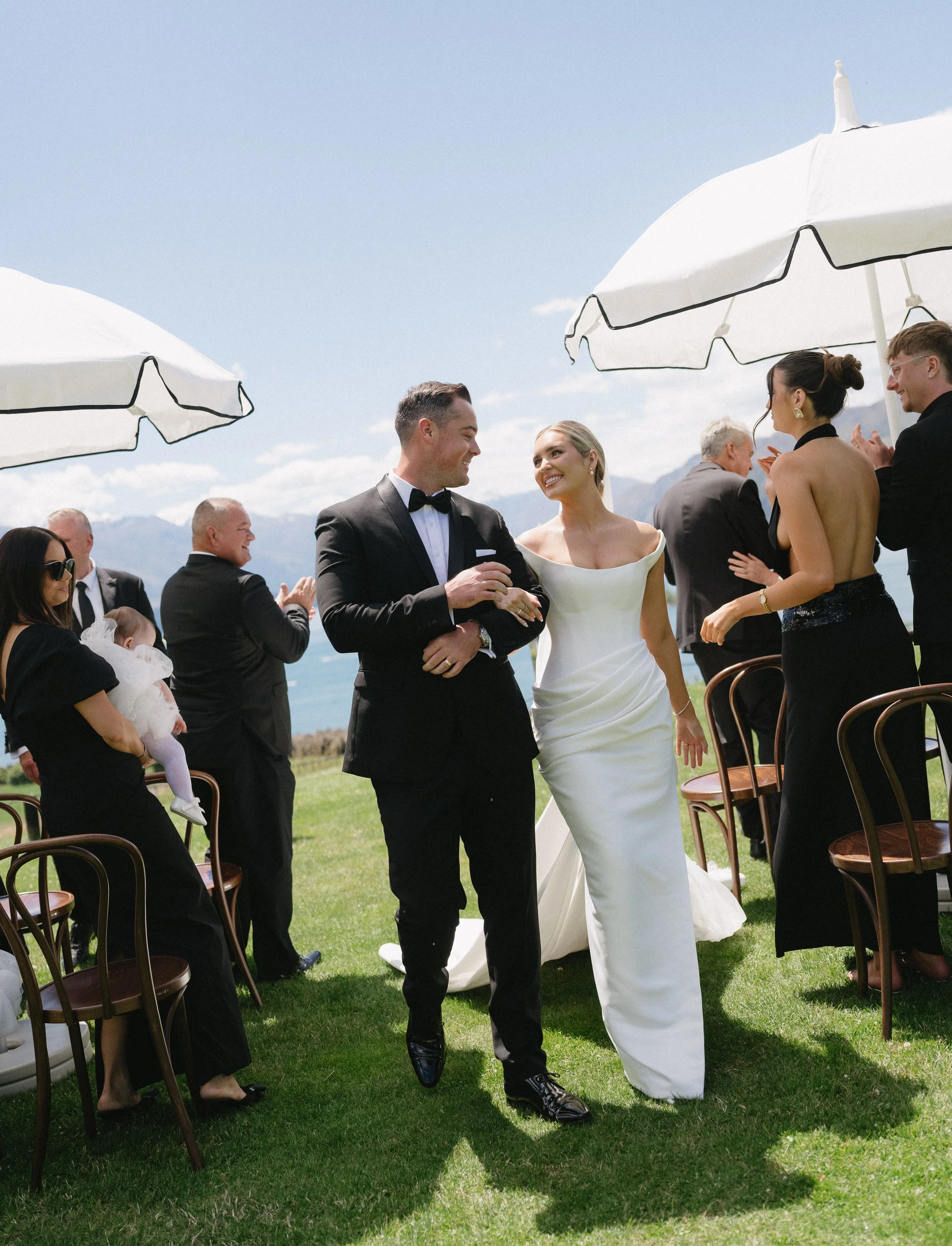 Bride and groom walking arm-in-arm at outdoor wedding reception under umbrellas, surrounded by guests clapping and celebrating on a sunny day.