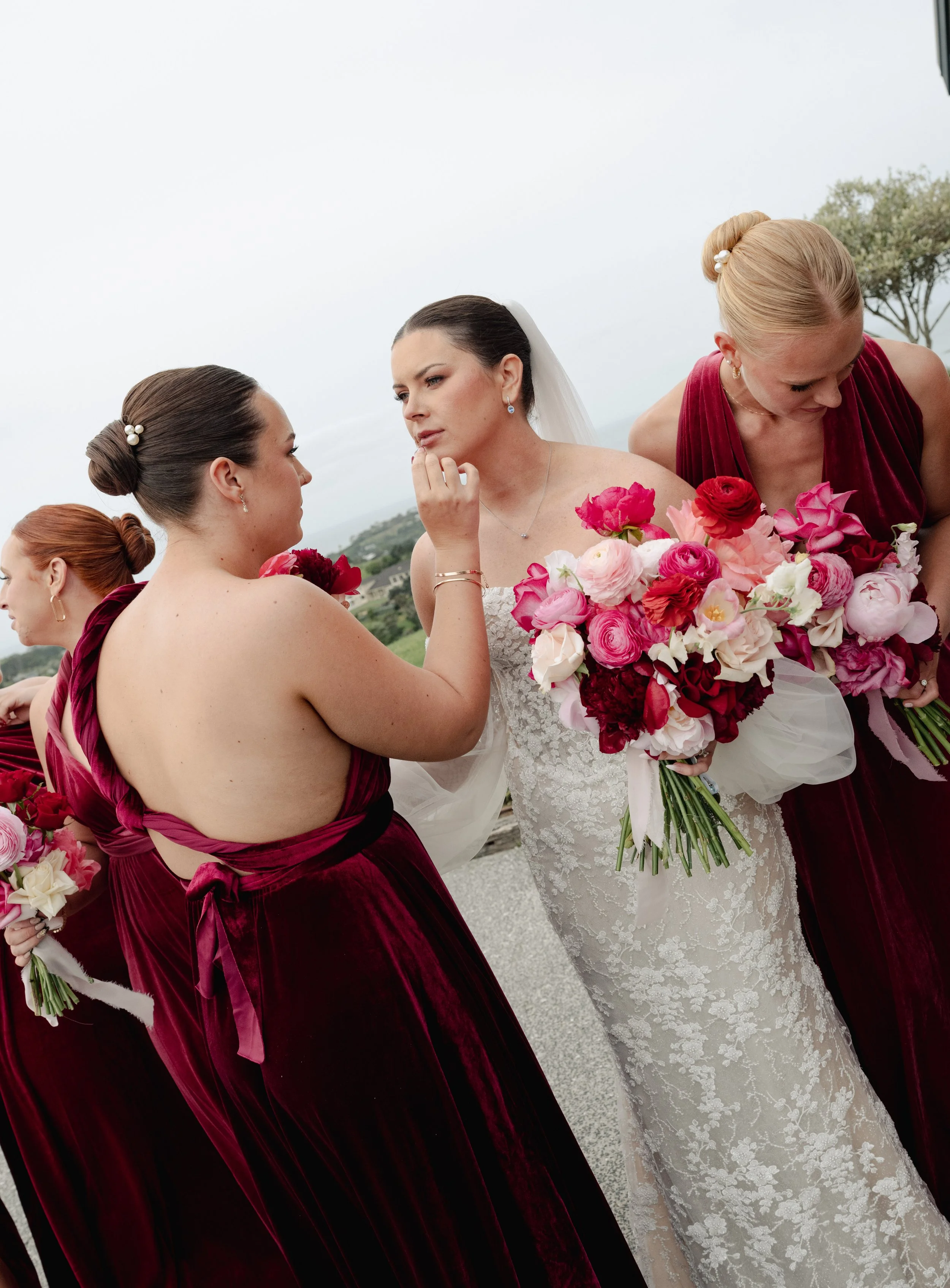 A bride in a white lace wedding dress holding a large bouquet of pink, red, and white flowers. Three bridesmaids in dark red velvet dresses with matching bouquets, helping the bride with her dress and makeup, outdoors on a cloudy day.