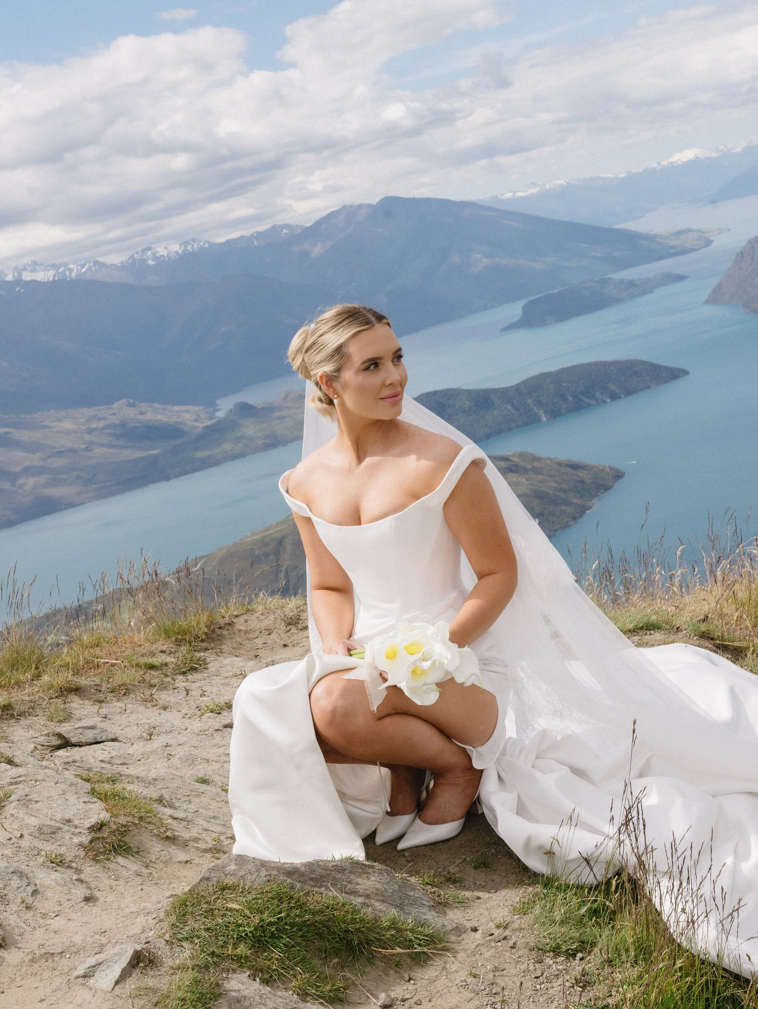 A woman in a wedding dress sitting on grass with a mountain and lake landscape in the background.