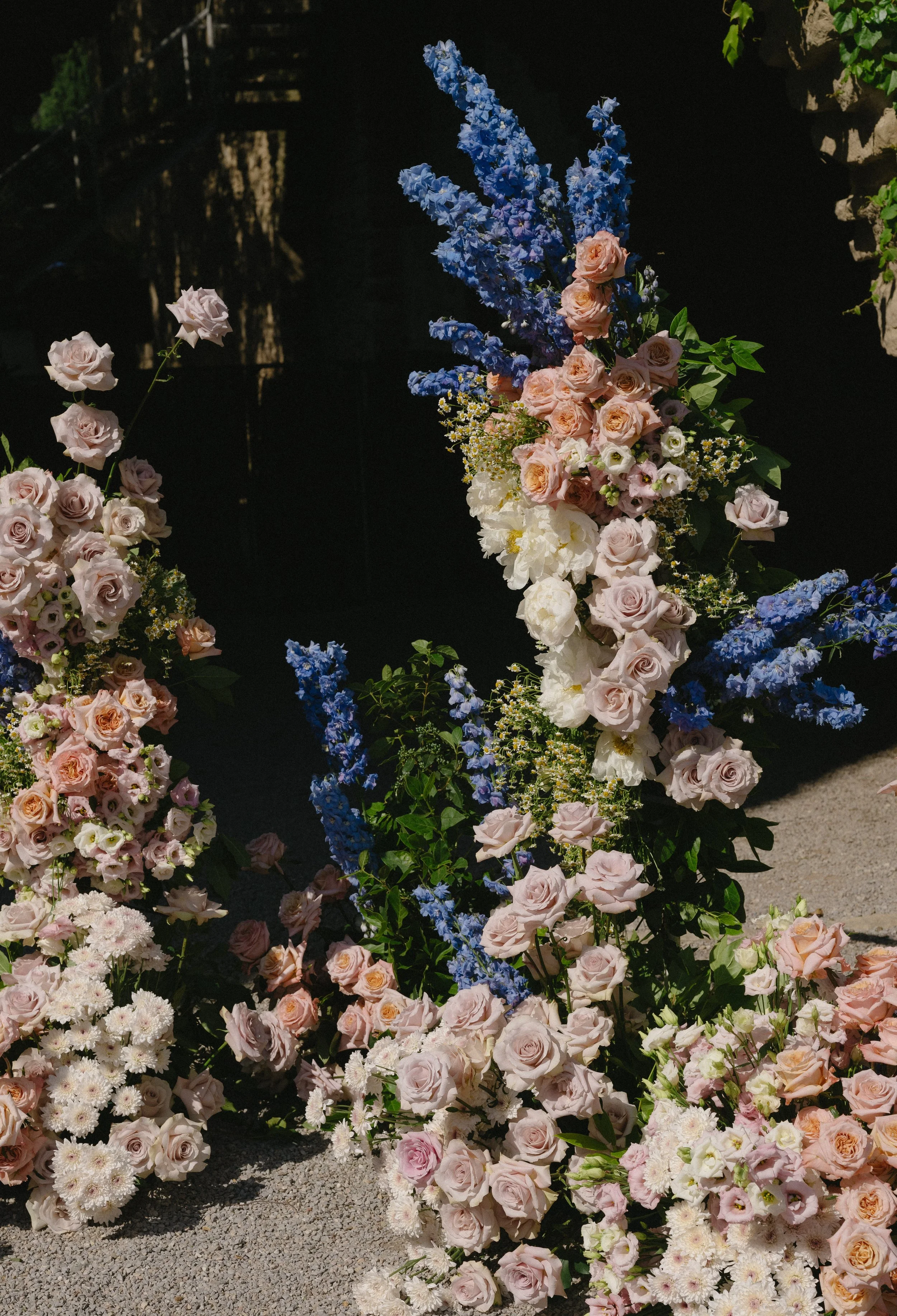 A variety of pastel-colored roses, delphiniums, and other flowers arranged in a floral display under sunlight.