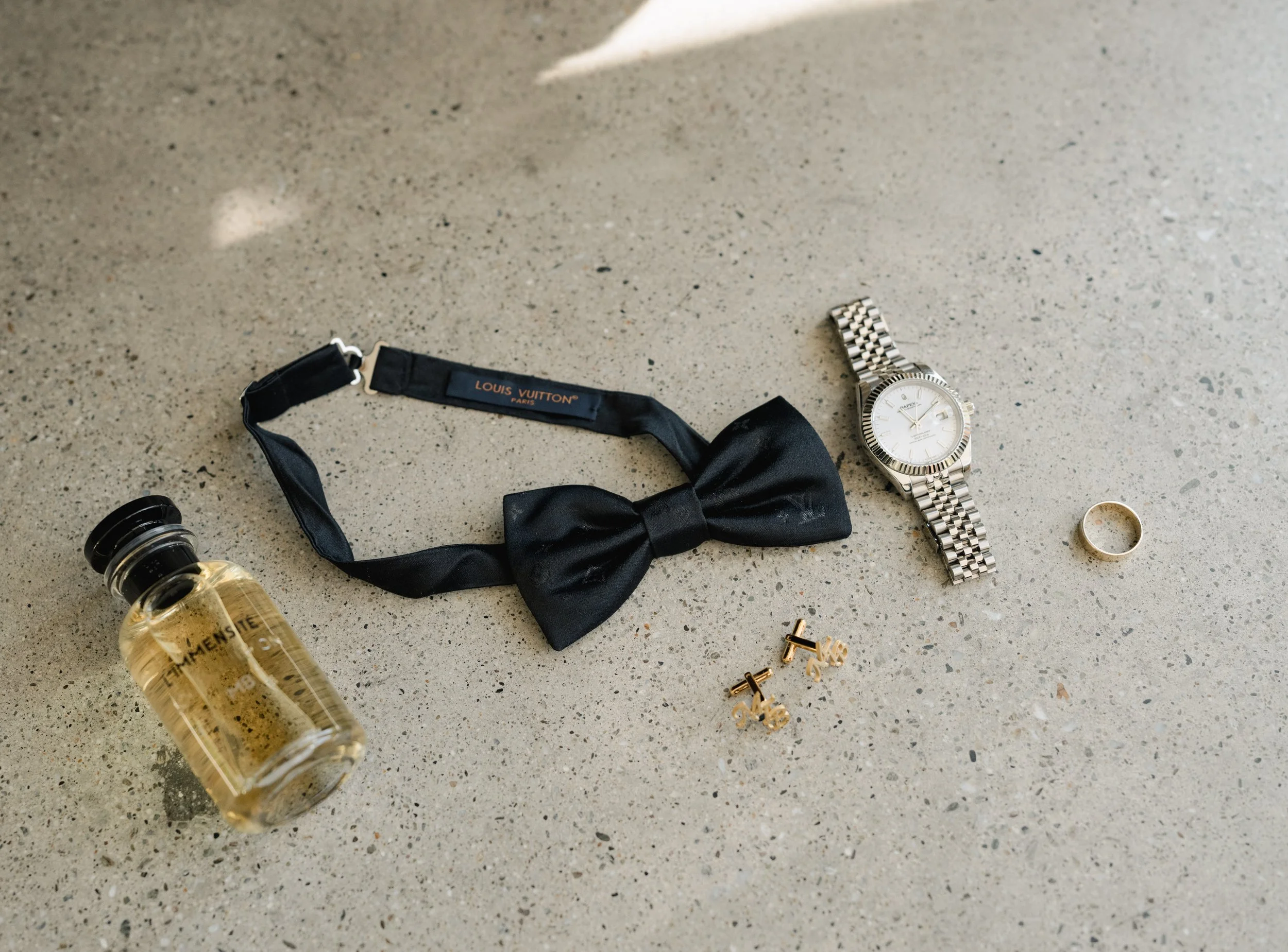 A collection of men's accessories on a concrete surface, including a bottle of cologne, a black Louis Vuitton bow tie with matching strap, a silver wristwatch, a gold wedding band, gold cufflinks, and a gold ring.