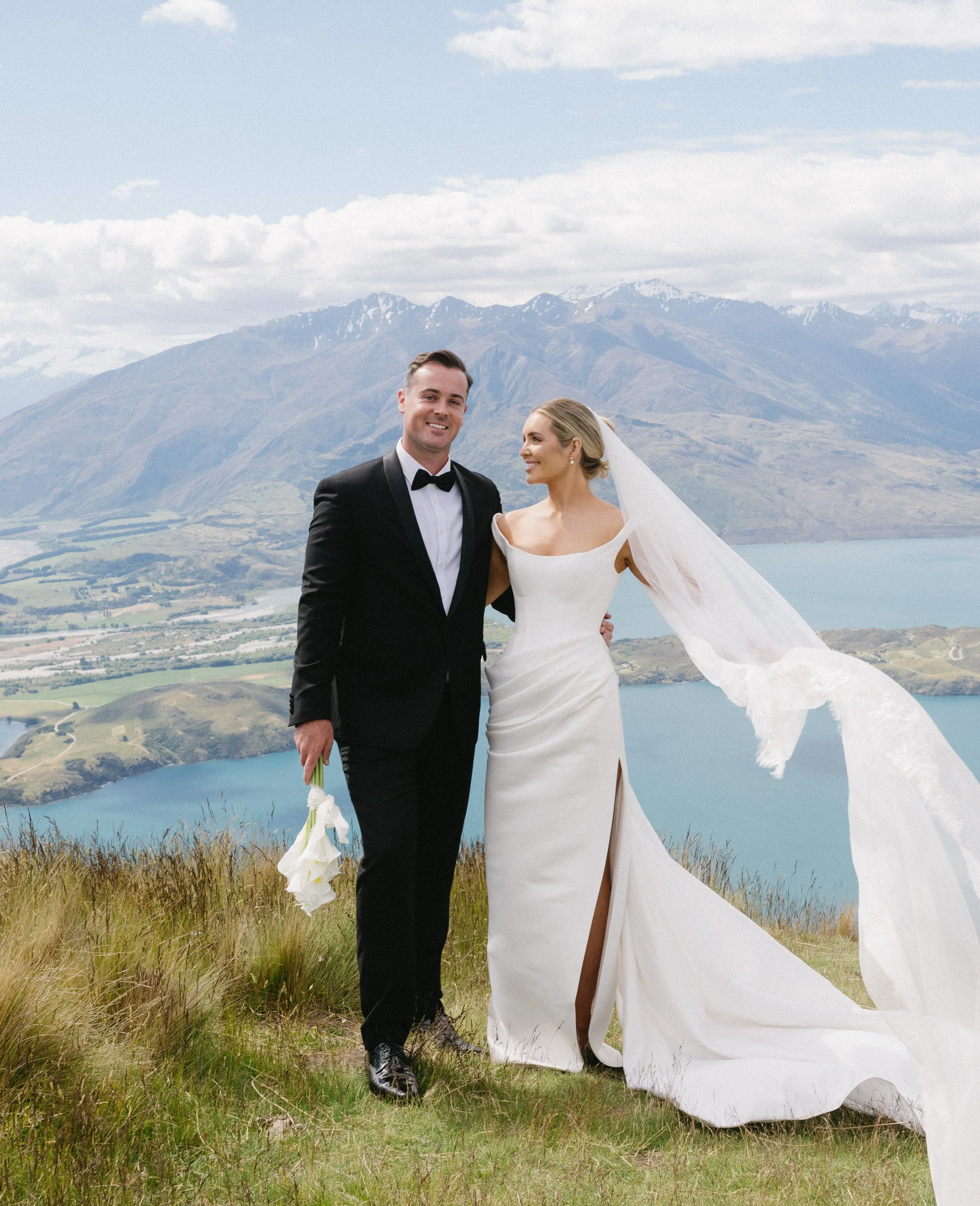 A bride and groom standing outdoors in front of a scenic mountain and lake backdrop, with the groom in a tuxedo and the bride in a white wedding gown.