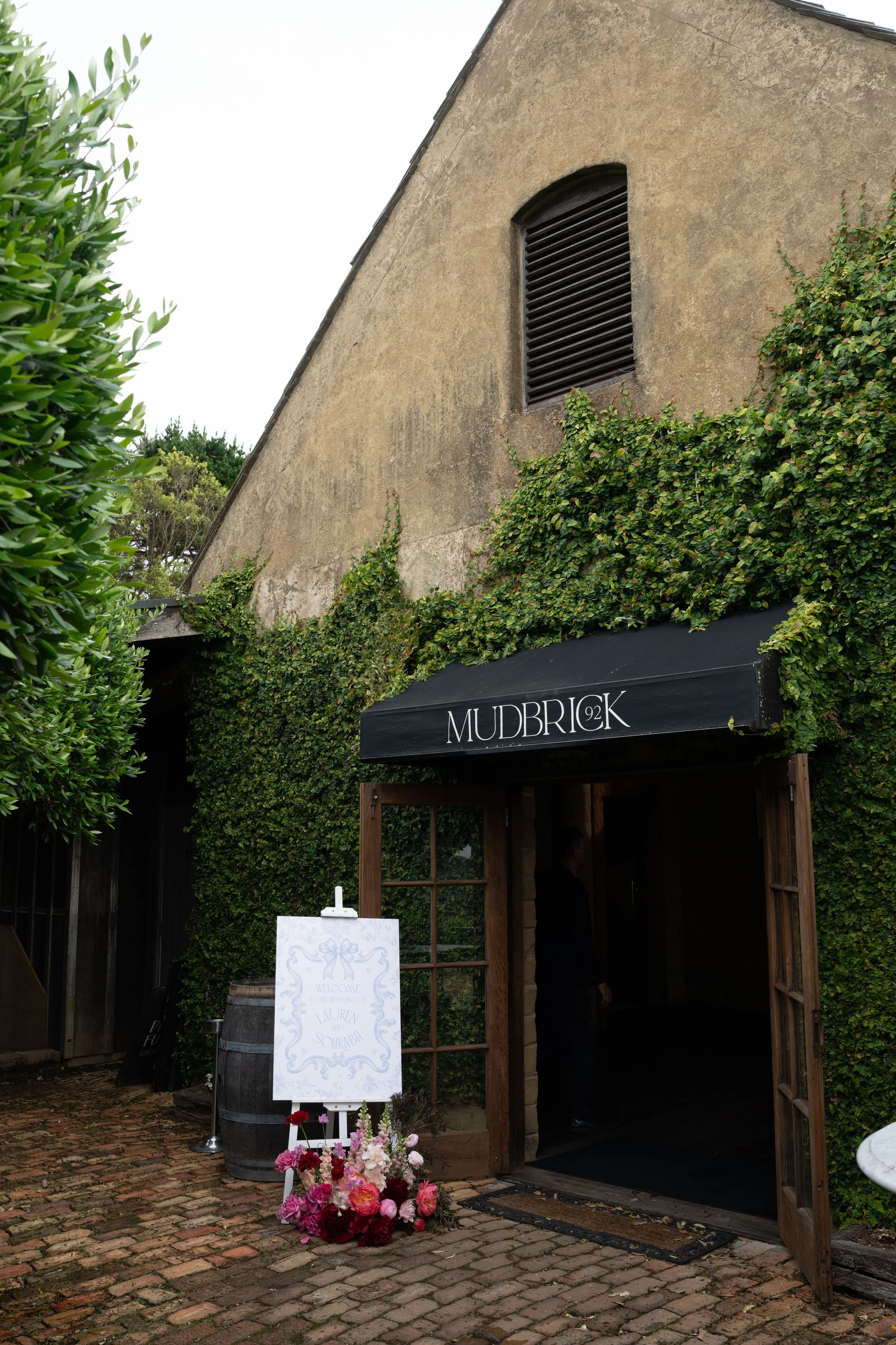 Entrance to a venue called Mudbrick, decorated for a wedding with a floral display and a welcome sign for Lauren and Shrub. The building has a stucco exterior with an arched window and is surrounded by greenery.
