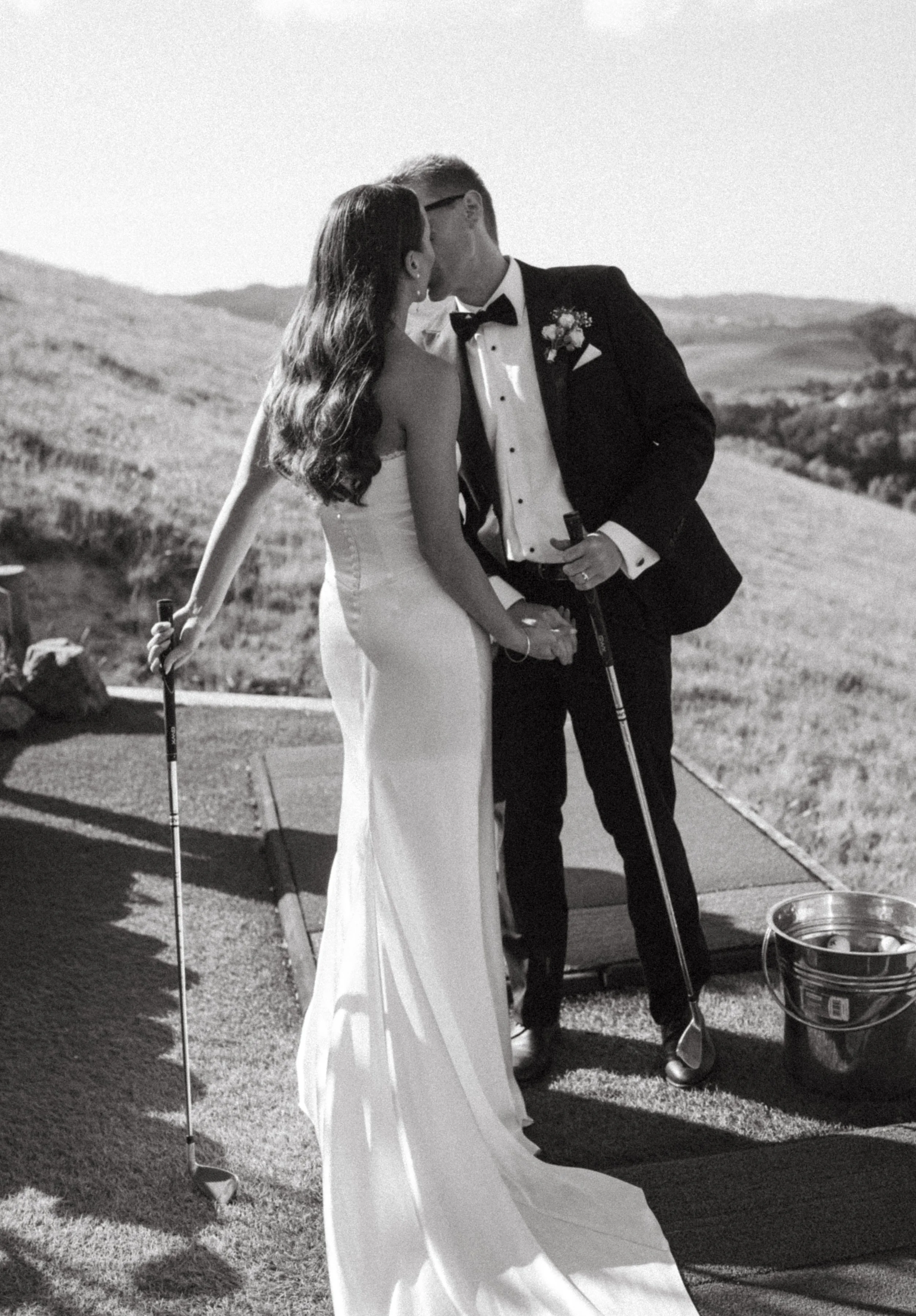 A couple dressed in wedding attire, the bride in a white gown and the groom in a tuxedo, sharing a kiss while holding golf clubs outdoors.