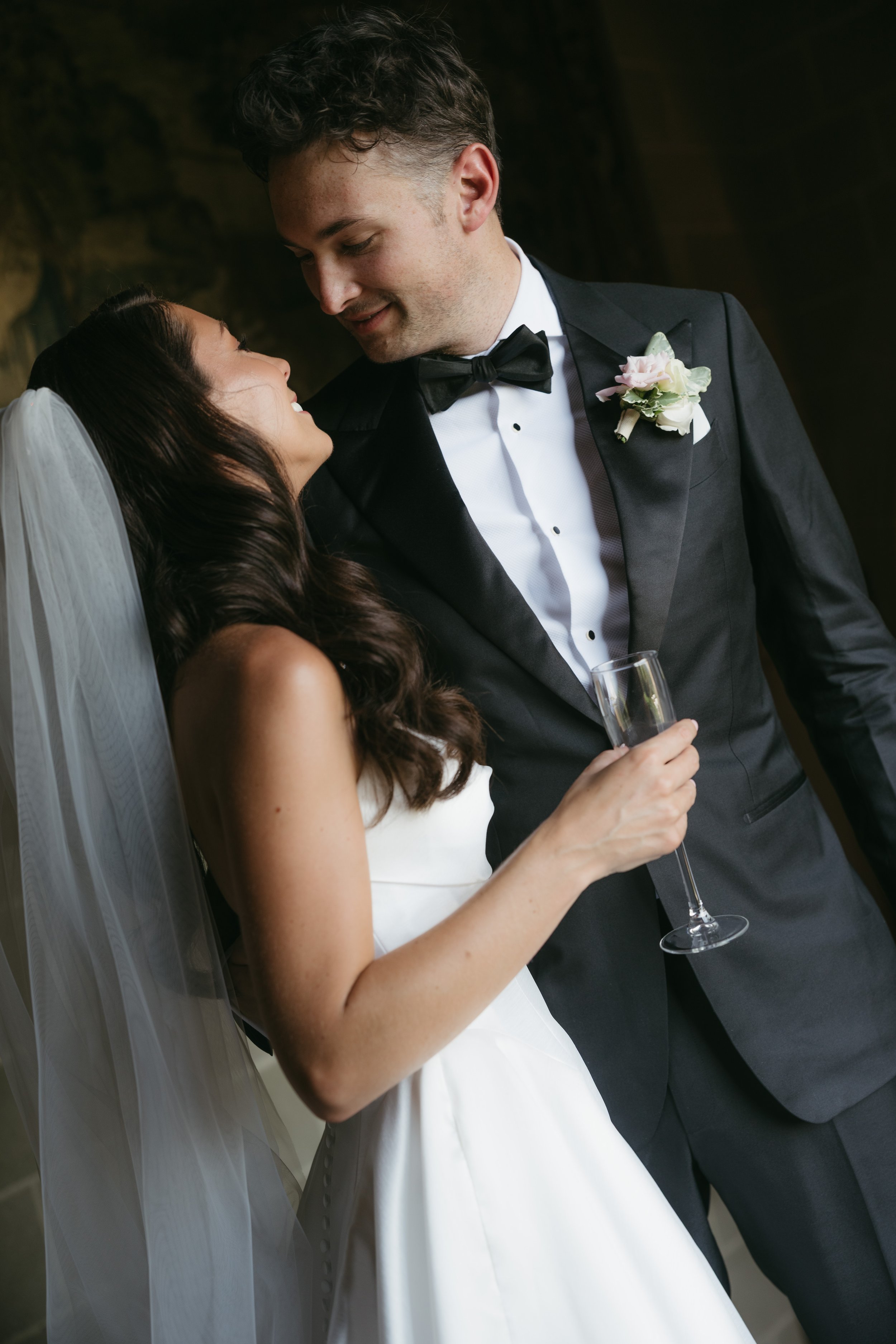 A bride and groom at their wedding, smiling and looking at each other, with the bride holding a champagne flute.