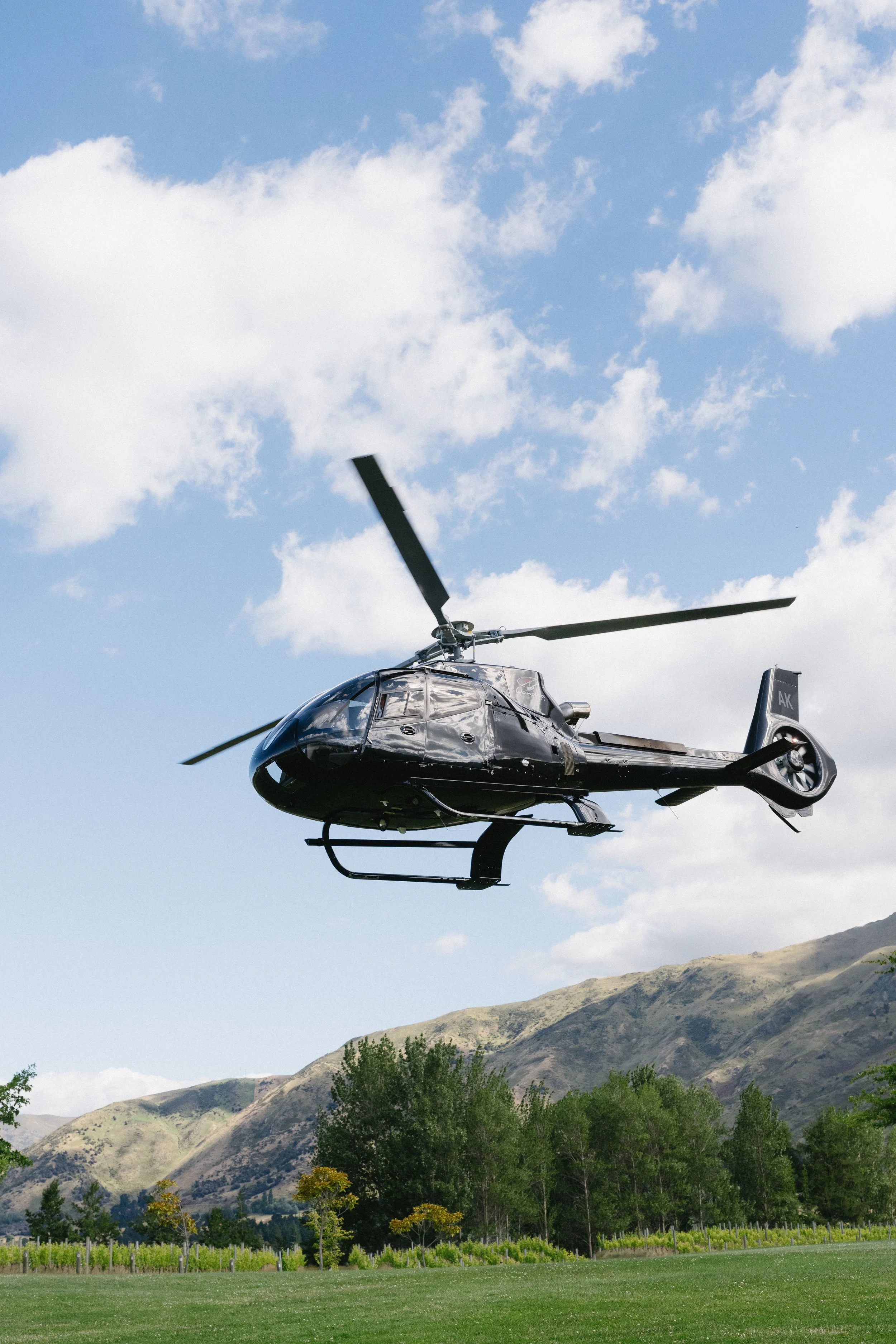 A black helicopter flying in the sky over a green landscape with trees and hills.