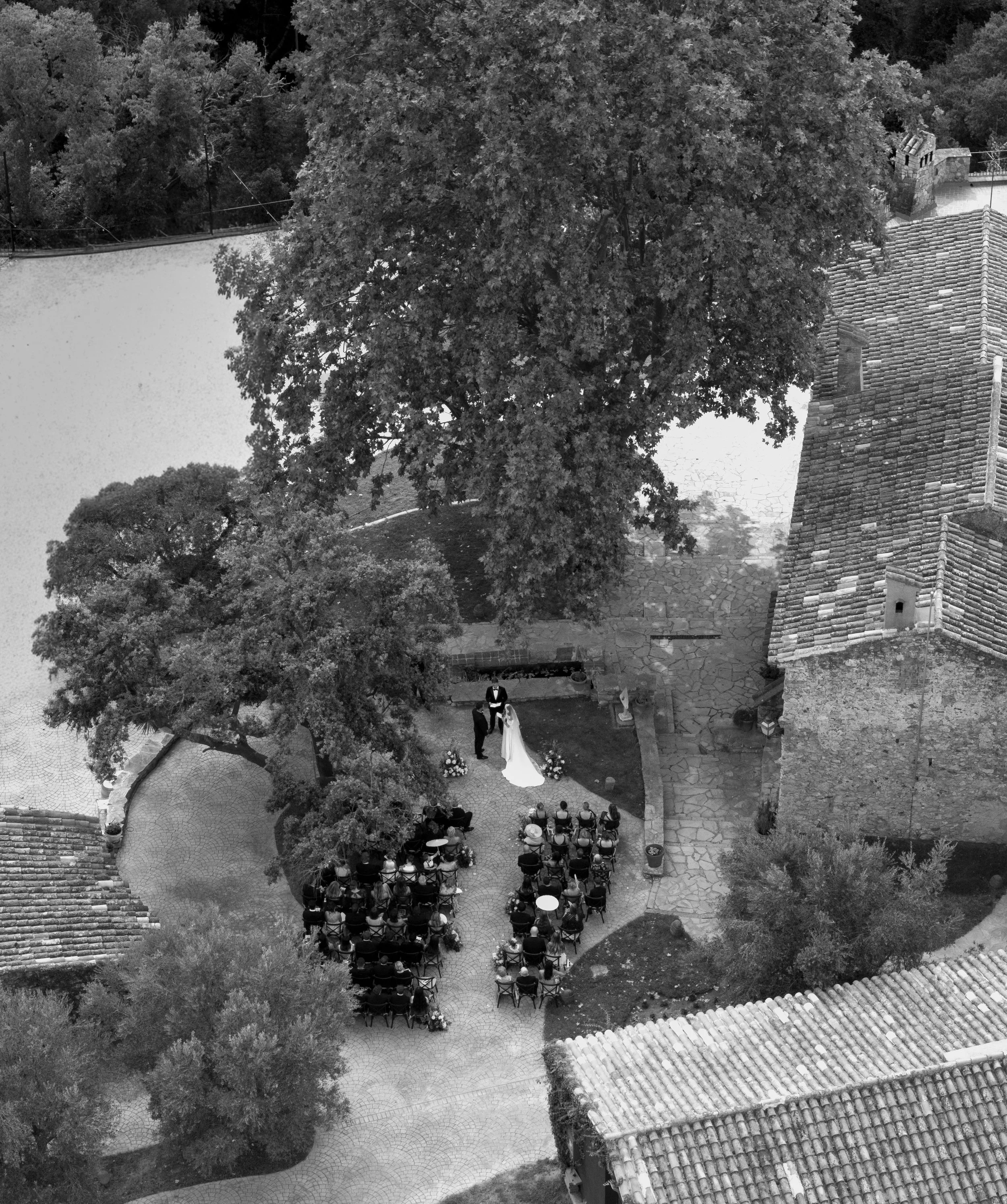 Black and white aerial view of a wedding ceremony in a courtyard surrounded by trees and brick buildings, with guests seated and a couple standing under an arch.