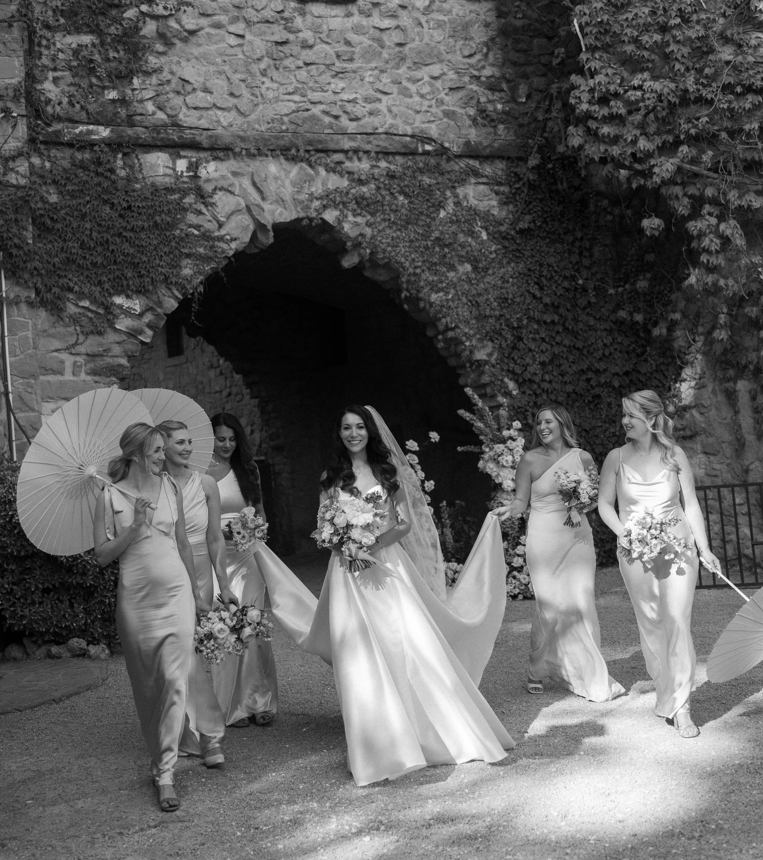 A bride in a wedding dress walking with five bridesmaids and holding bouquets, outdoors near a stone archway covered with ivy, in black and white.