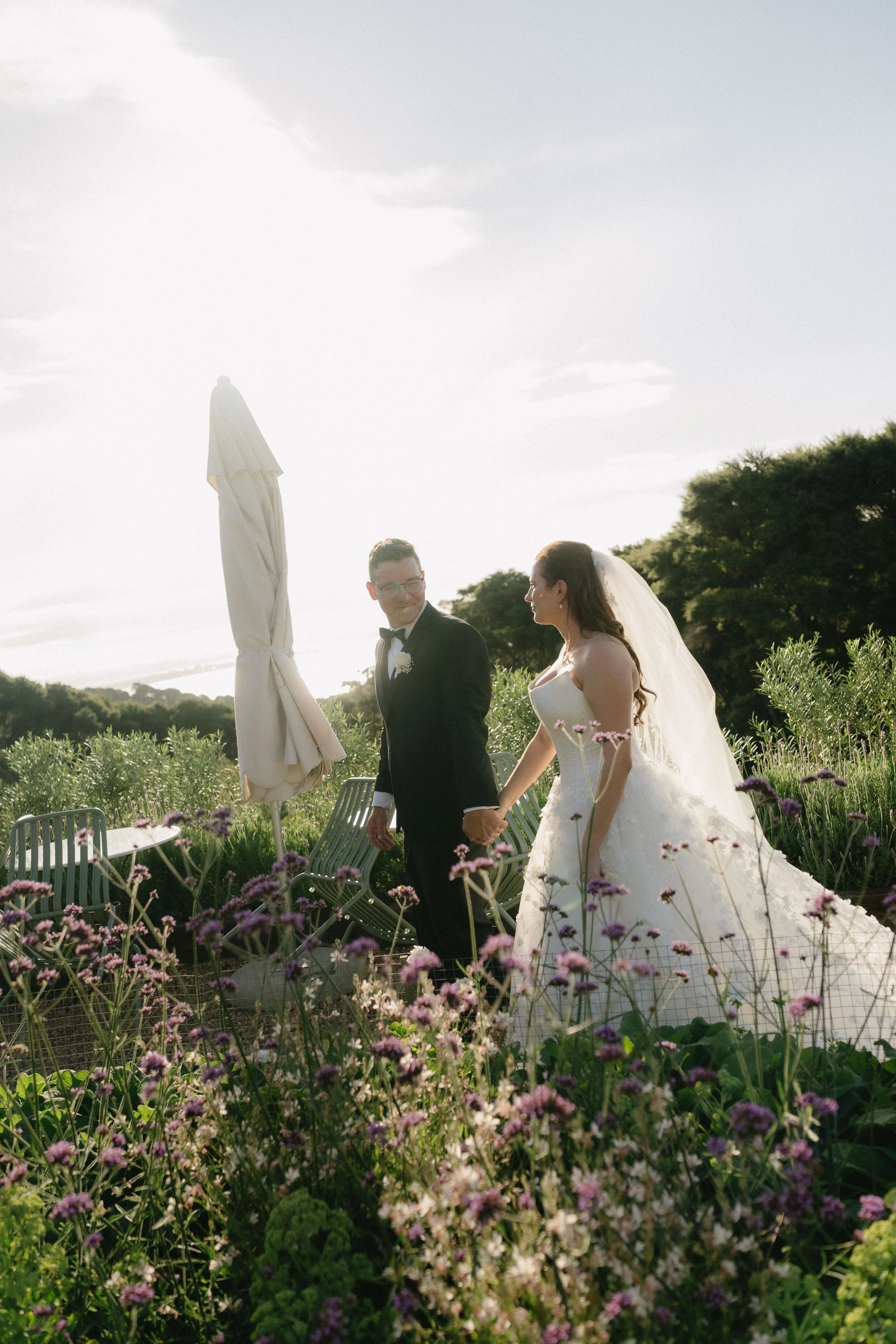 A bride and groom walking hand in hand through a garden with purple flowers, holding hands, with a cloudy sky and trees in the background.