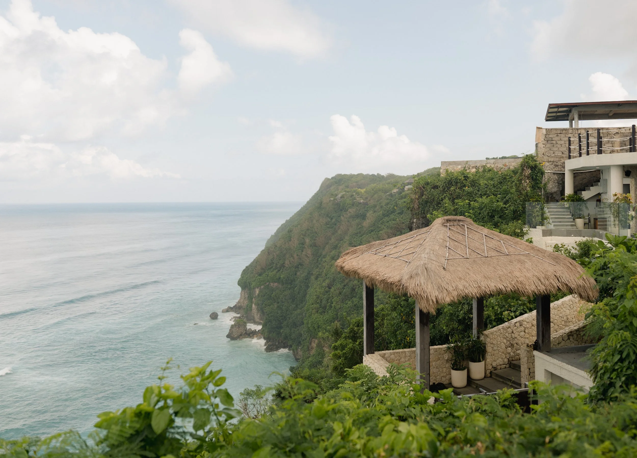 Ocean view from a cliffside resort with a thatched roof cabana and modern building with greenery.