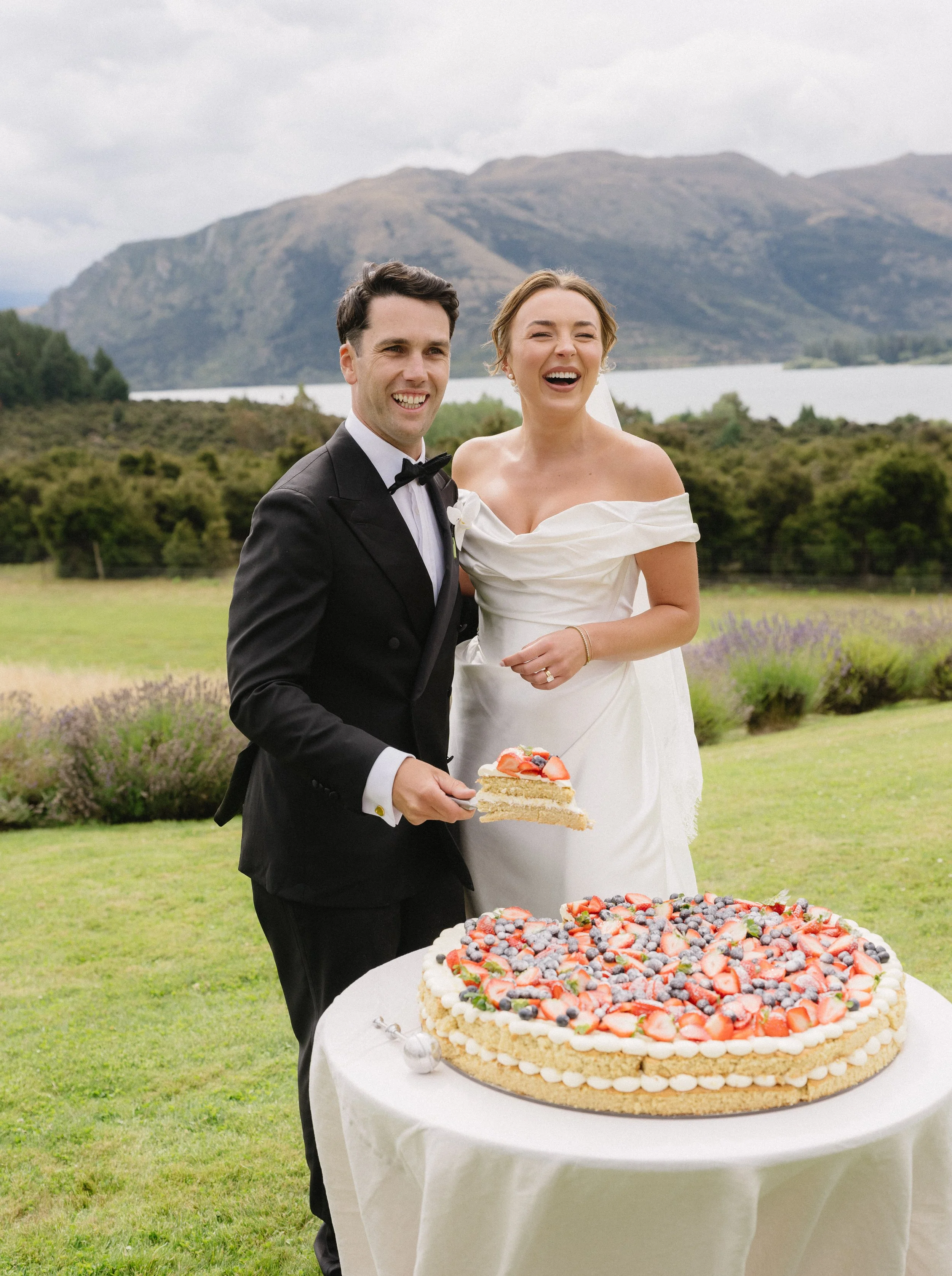 A happy bride and groom standing outdoors with mountains and a lake in the background, holding a piece of cake near a large decorated cake on a table.