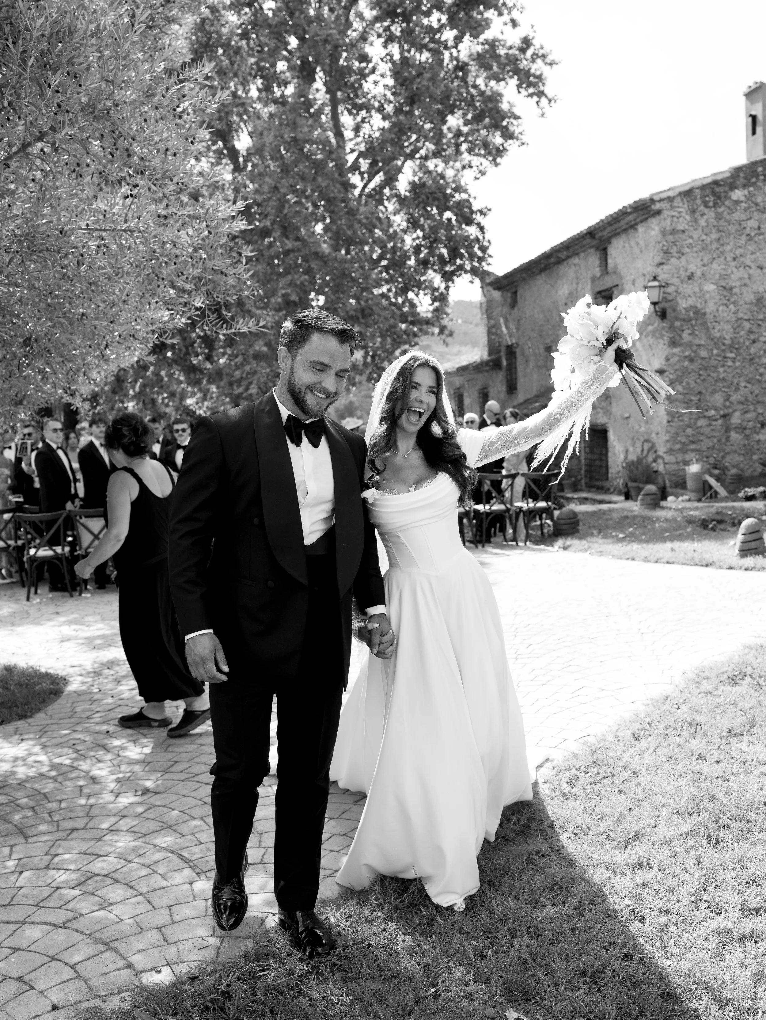 Black and white photo of a bride and groom holding hands and smiling, walking outdoors during a wedding celebration. The bride is wearing a wedding dress and veil, and the groom is in a tuxedo. Guests can be seen in the background, along with rustic 