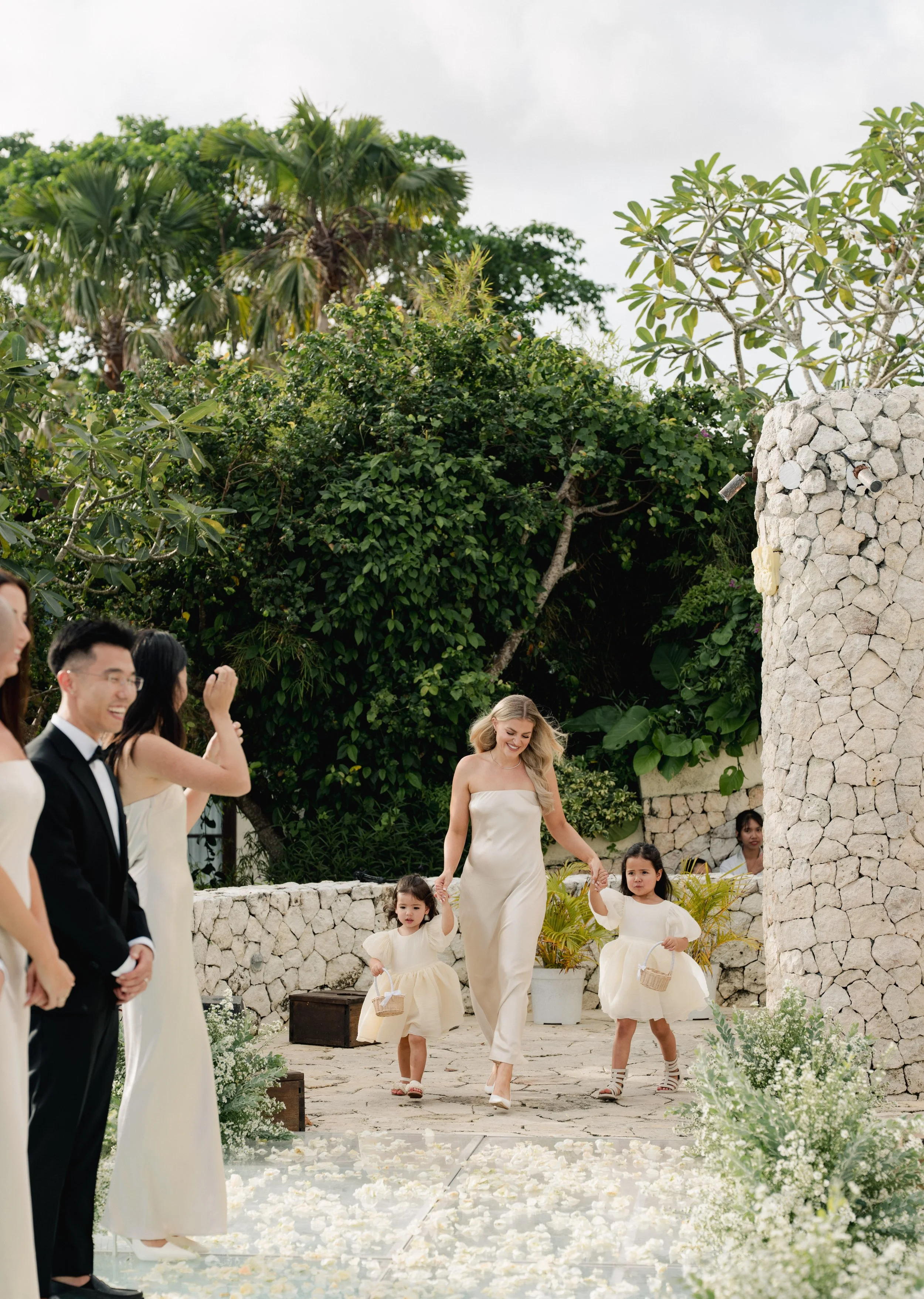 Women and children at a wedding ceremony outdoors, with lush greenery and stone structures in the background.