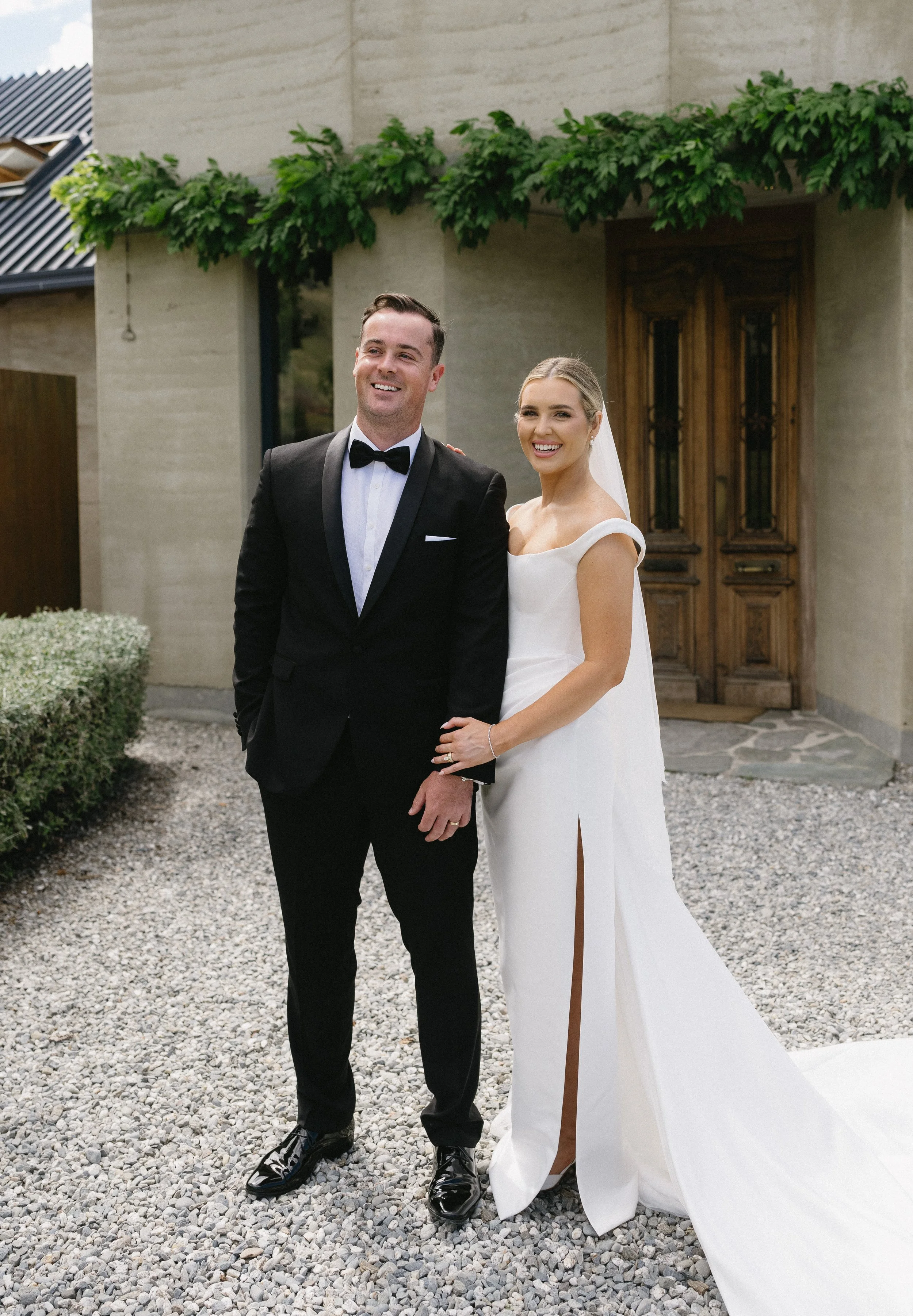 A smiling bride and groom dressed in wedding attire, standing outdoors on a gravel pathway in front of a building with greenery.