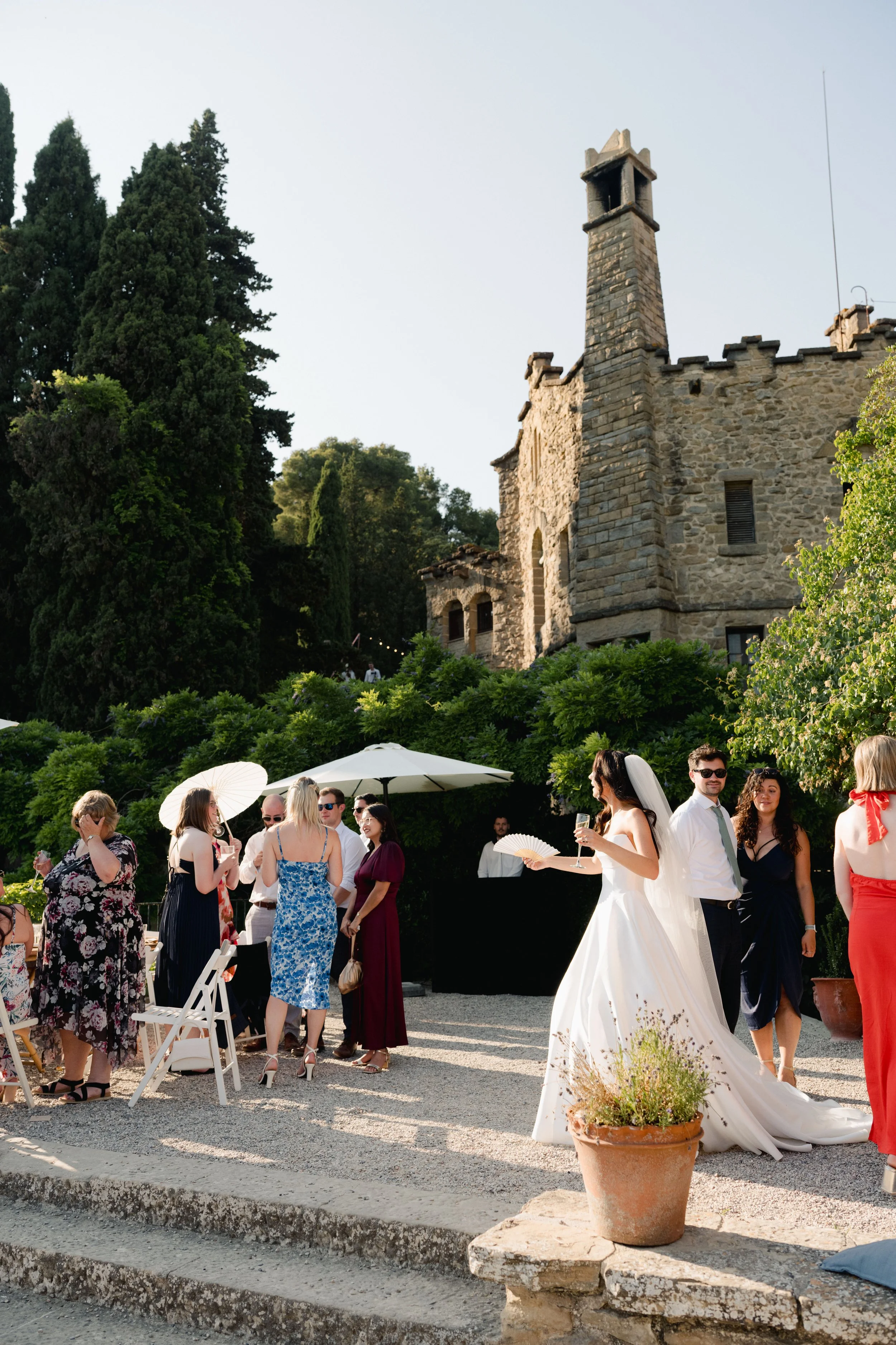 Outdoor wedding reception with guests socializing near a historic stone building, a bride in a white wedding dress with a veil holding a fan, surrounded by guests in formal attire, some under umbrellas, potted plants and greenery, in the late afterno