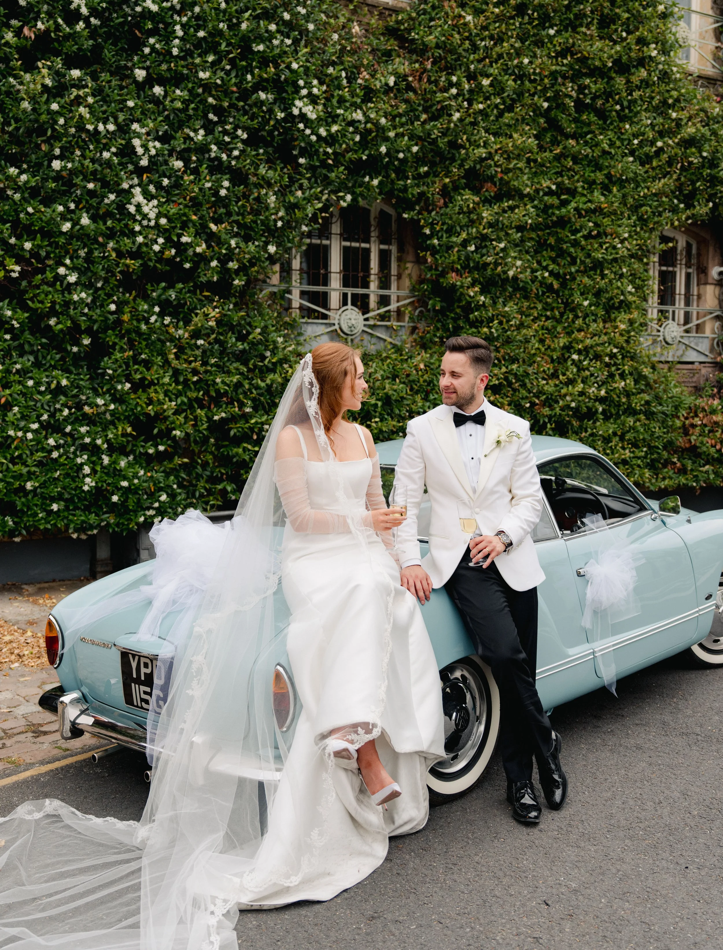 A bride and groom sitting on a vintage blue car decorated for a wedding, holding glasses of champagne, in front of a lush green hedge and a building.