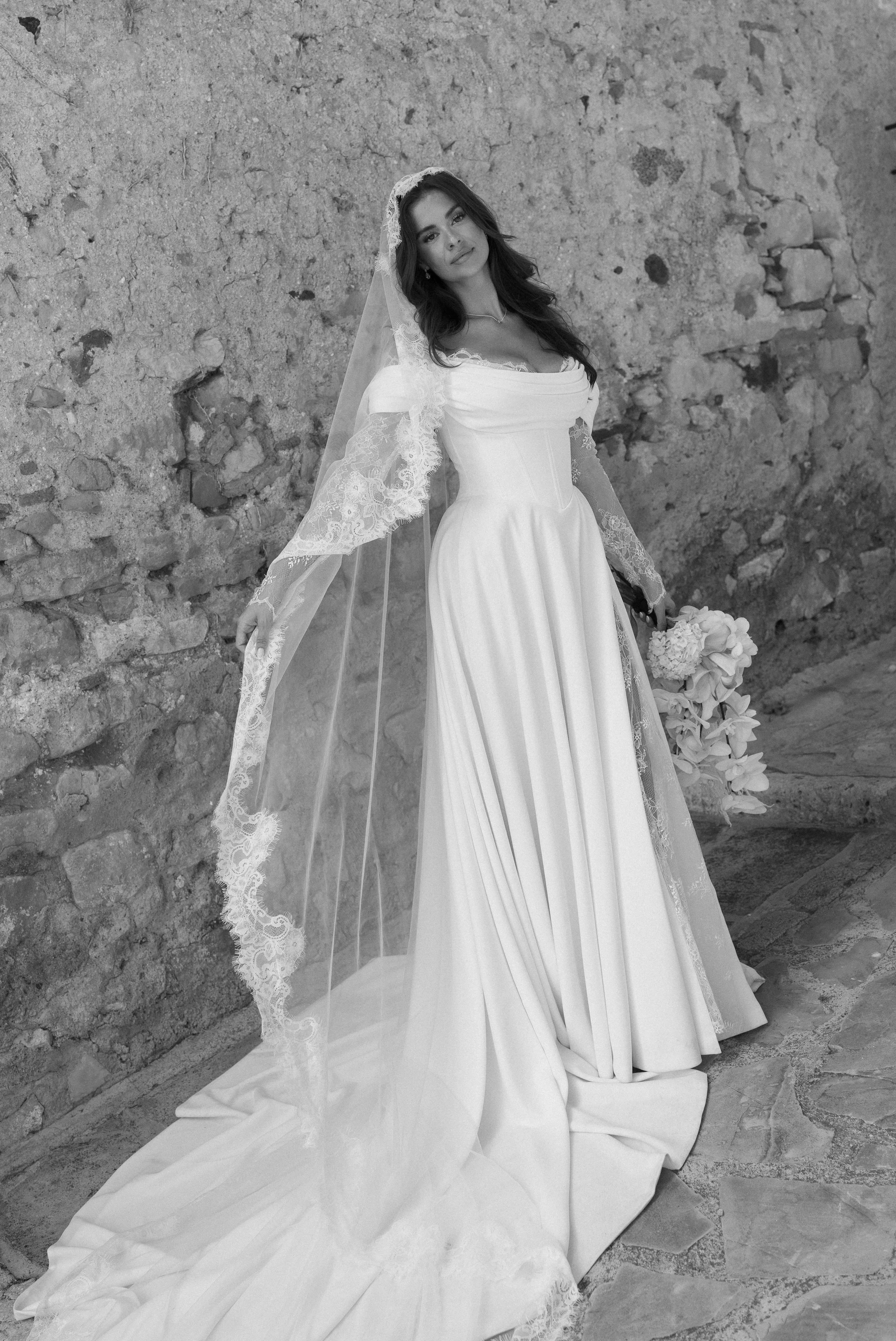 A woman in a wedding dress holding a bouquet of flowers, standing against a stone wall.
