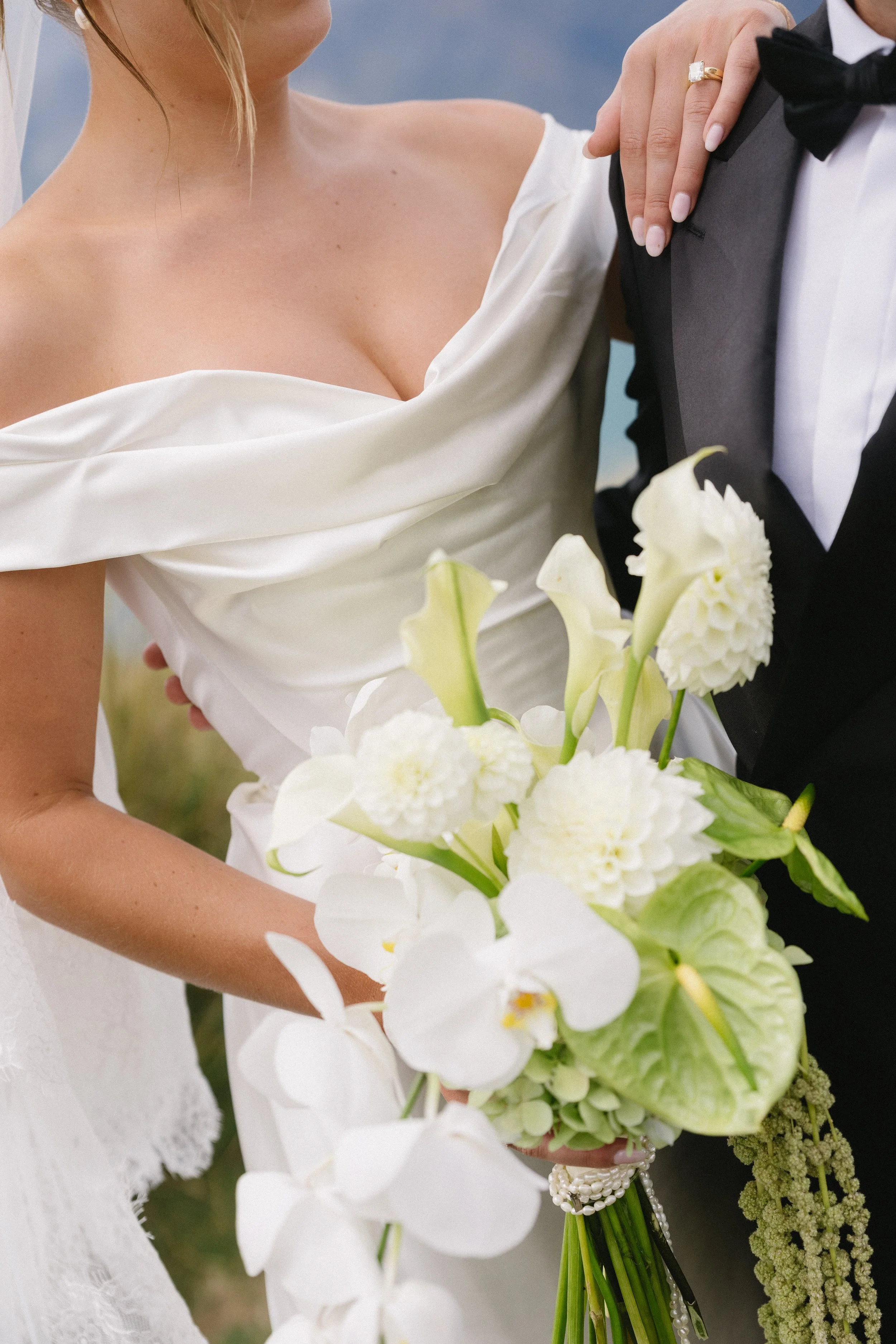 A bride and groom standing together, with the bride holding a large bouquet of white flowers including orchids and calla lilies, and the bride wearing an off-shoulder wedding dress while the groom is in a black tuxedo with a bow tie.