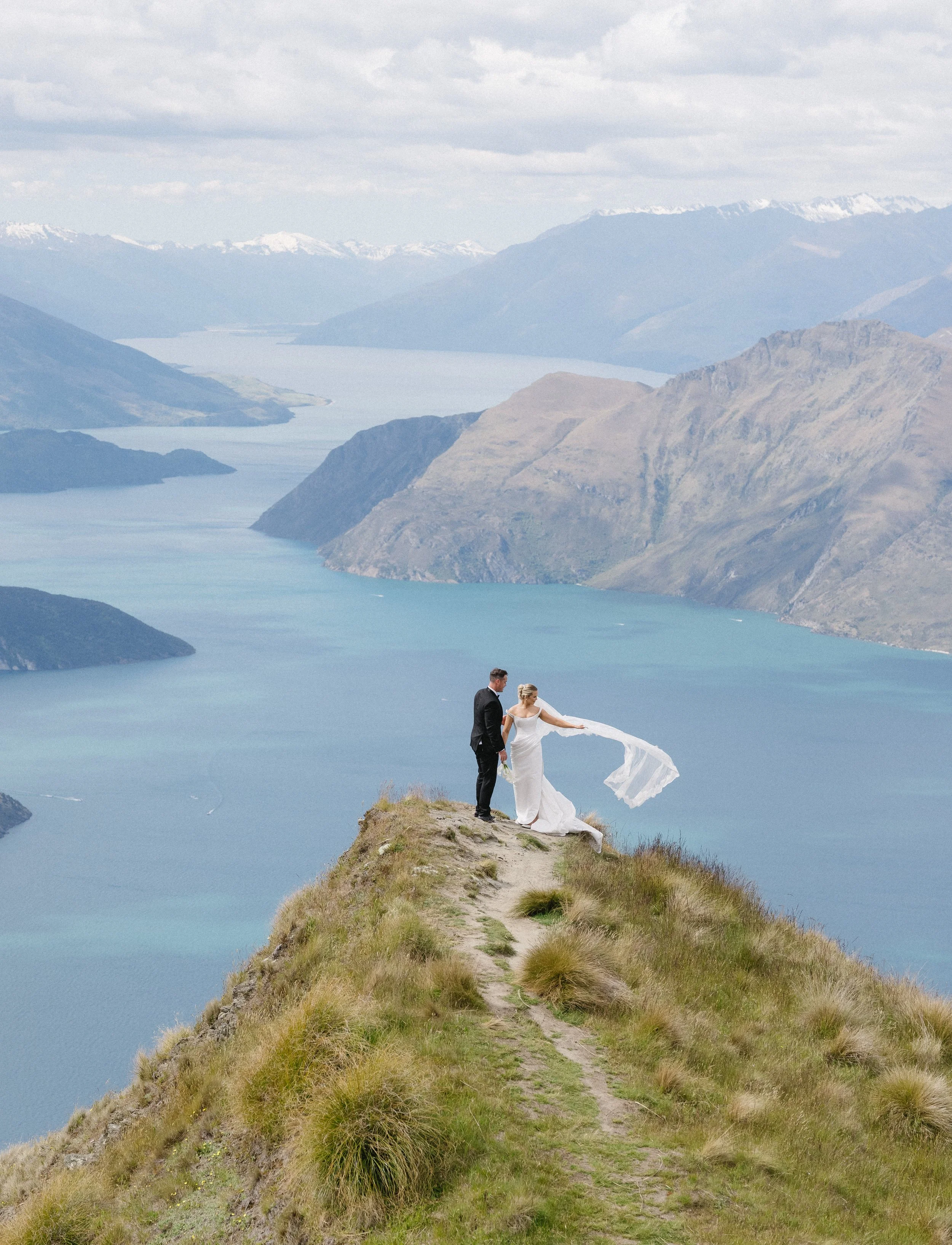 Bride and groom standing on a grassy mountain peak with a scenic view of a lake and mountains in the background.