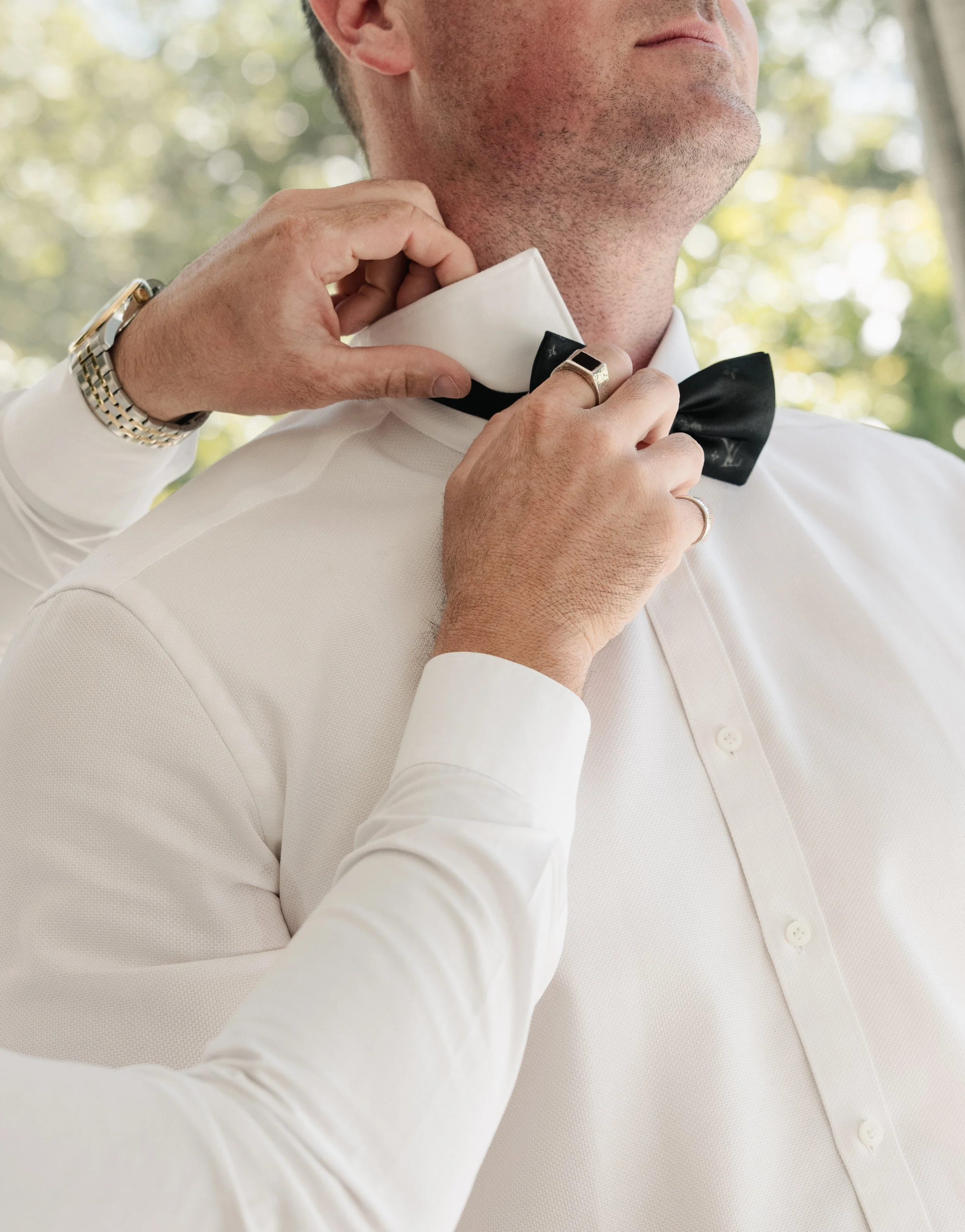 A person helping another man adjust a black bow tie on his white dress shirt, with a background of blurred outdoor greenery.