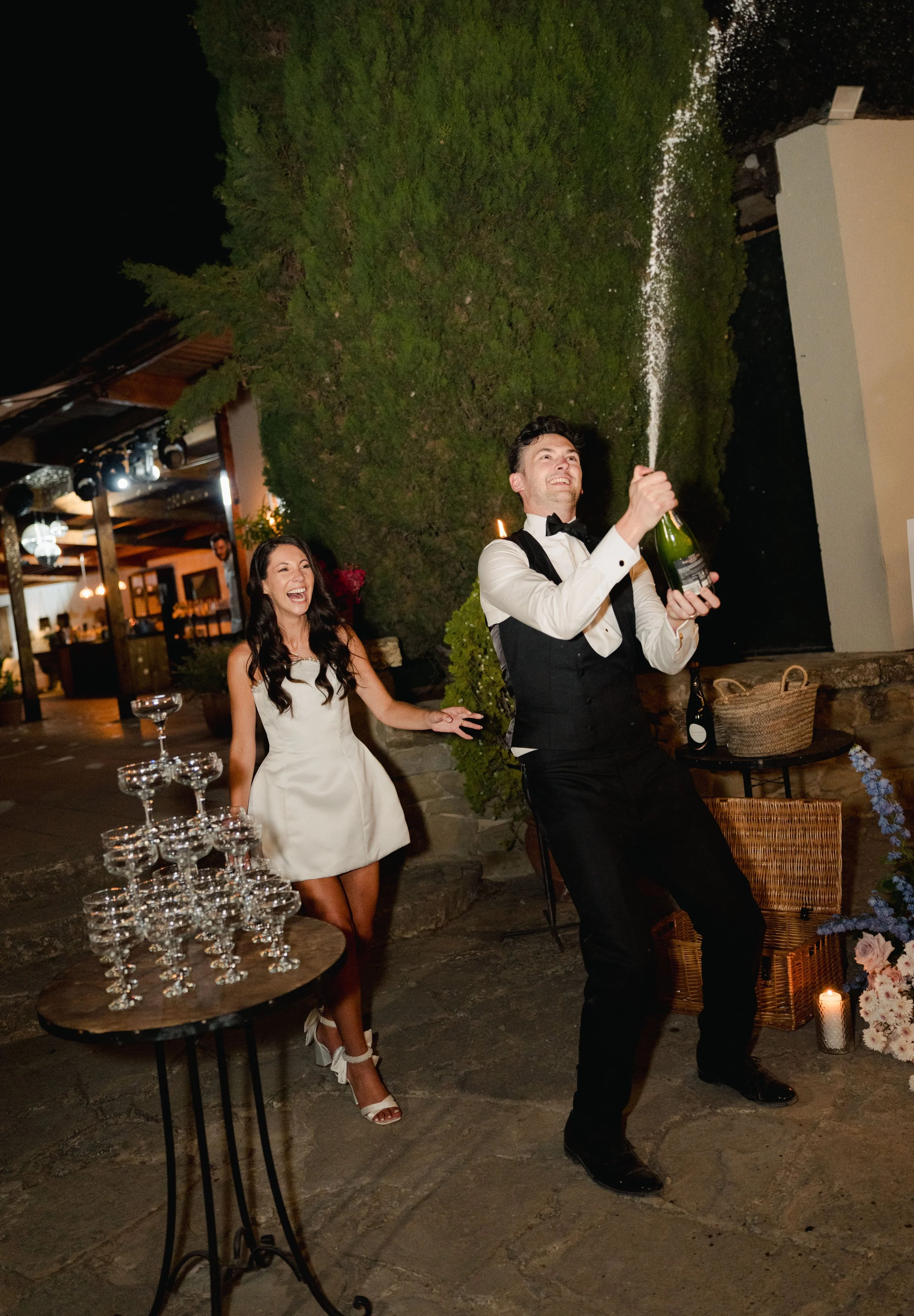 Couple celebrating, with the man opening a champagne bottle and the woman laughing, indoors at a wedding reception with champagne glasses on a table.