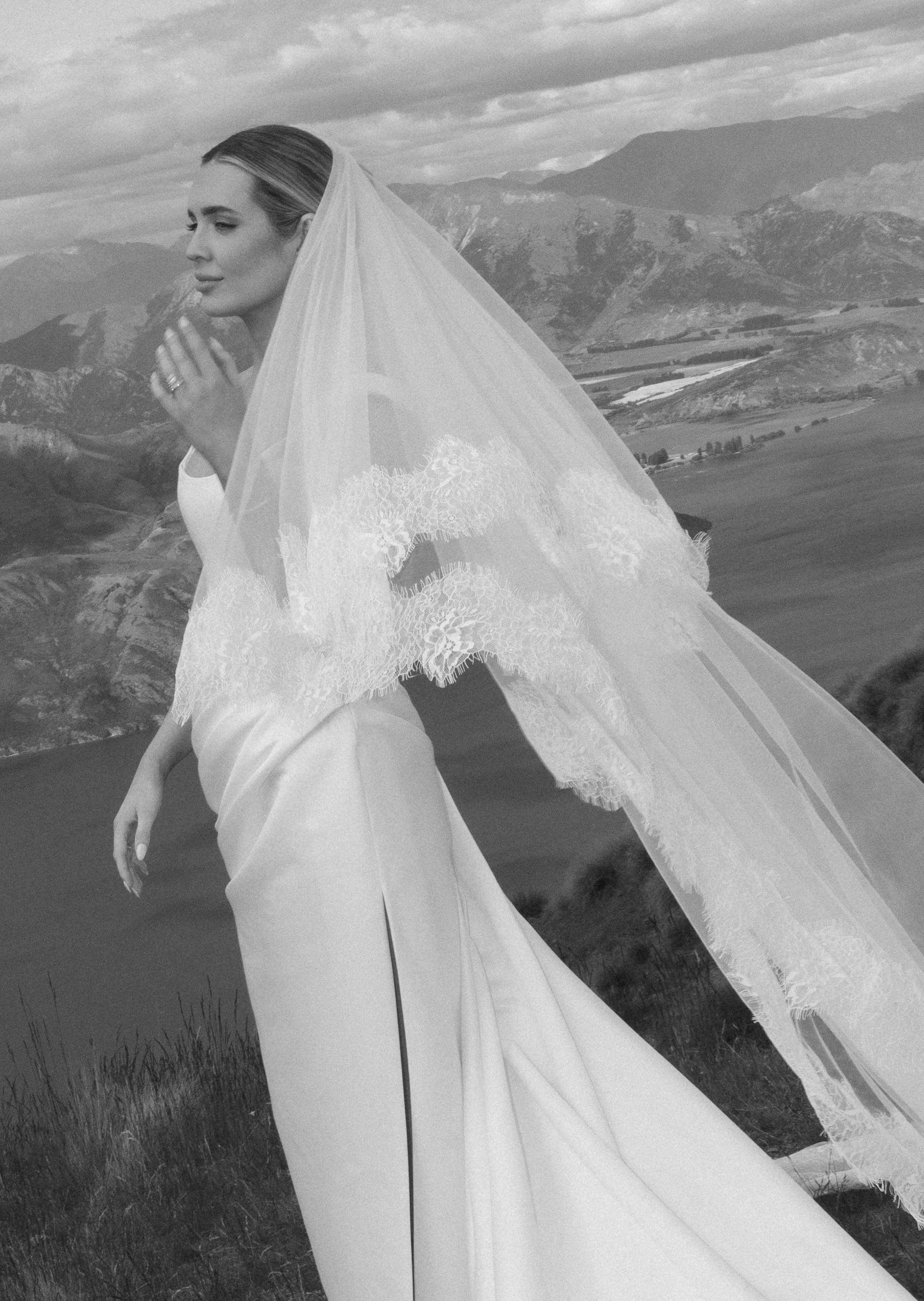A bride in a wedding dress and veil standing outdoors with mountains and a cloudy sky in the background.