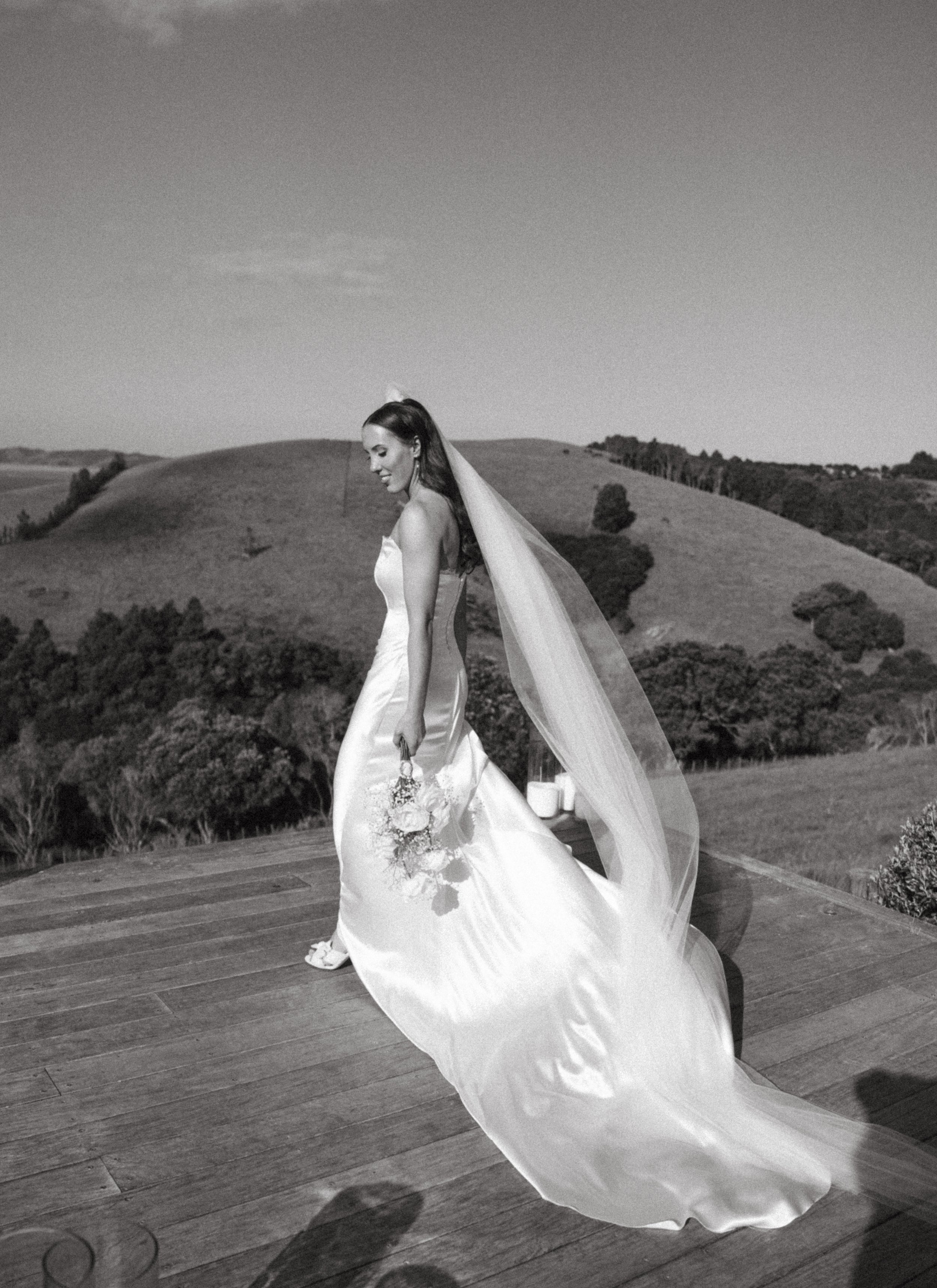 A bride in a wedding dress, holding a bouquet, standing on a wooden outdoor deck with rolling hills and trees in the background.