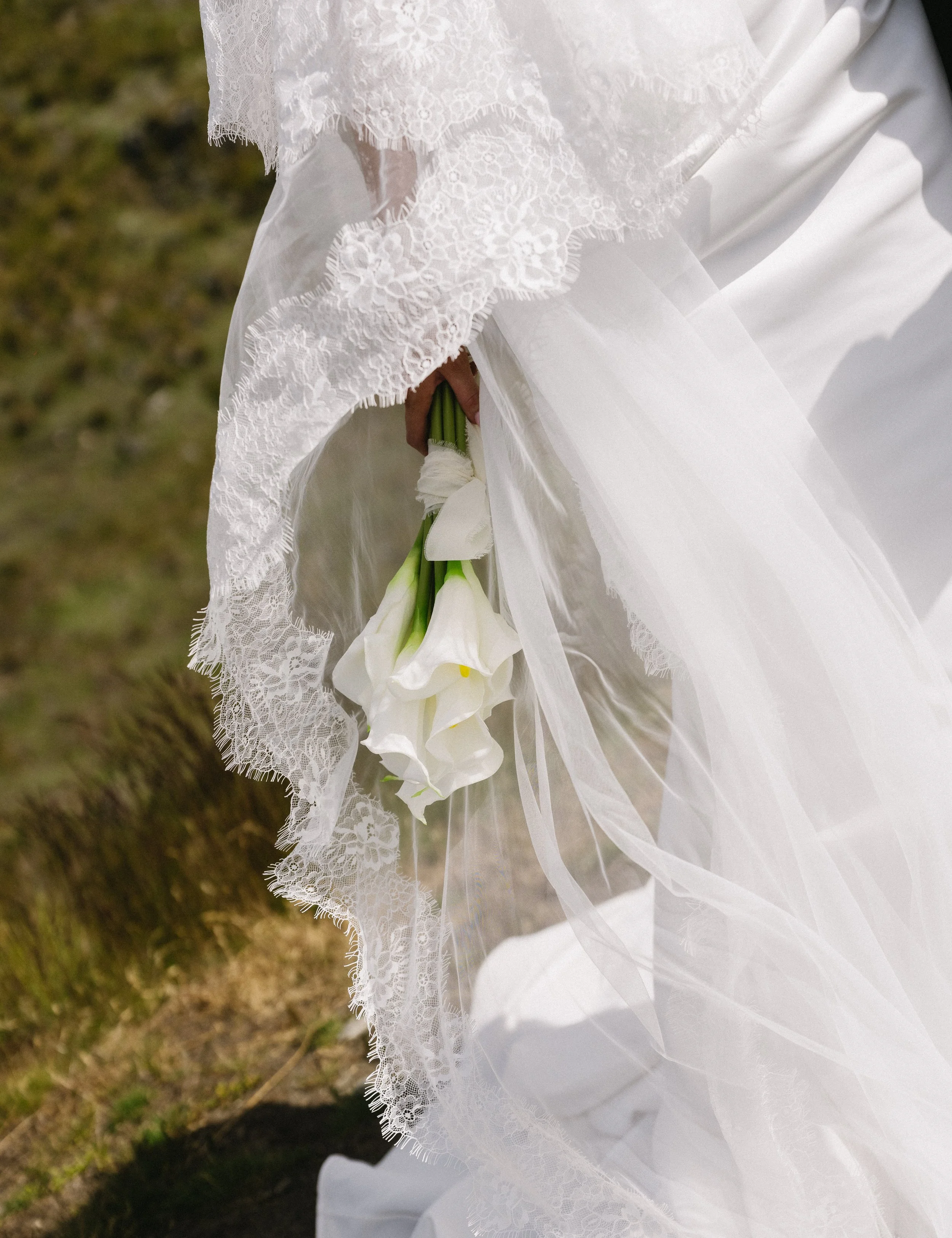 A bride holding a bouquet of white calla lilies, wearing a lace and tulle wedding dress.
