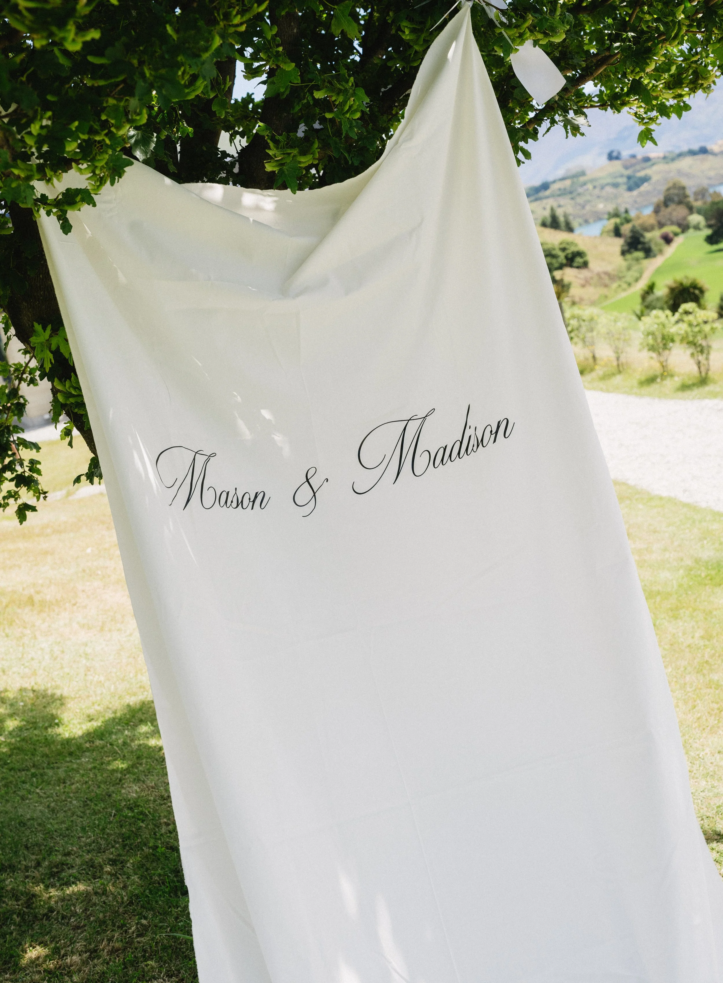 White fabric with elegant black script reading 'Mason & Madison,' hanging from a tree in an outdoor setting with green grass, trees, and rolling hills in the background.