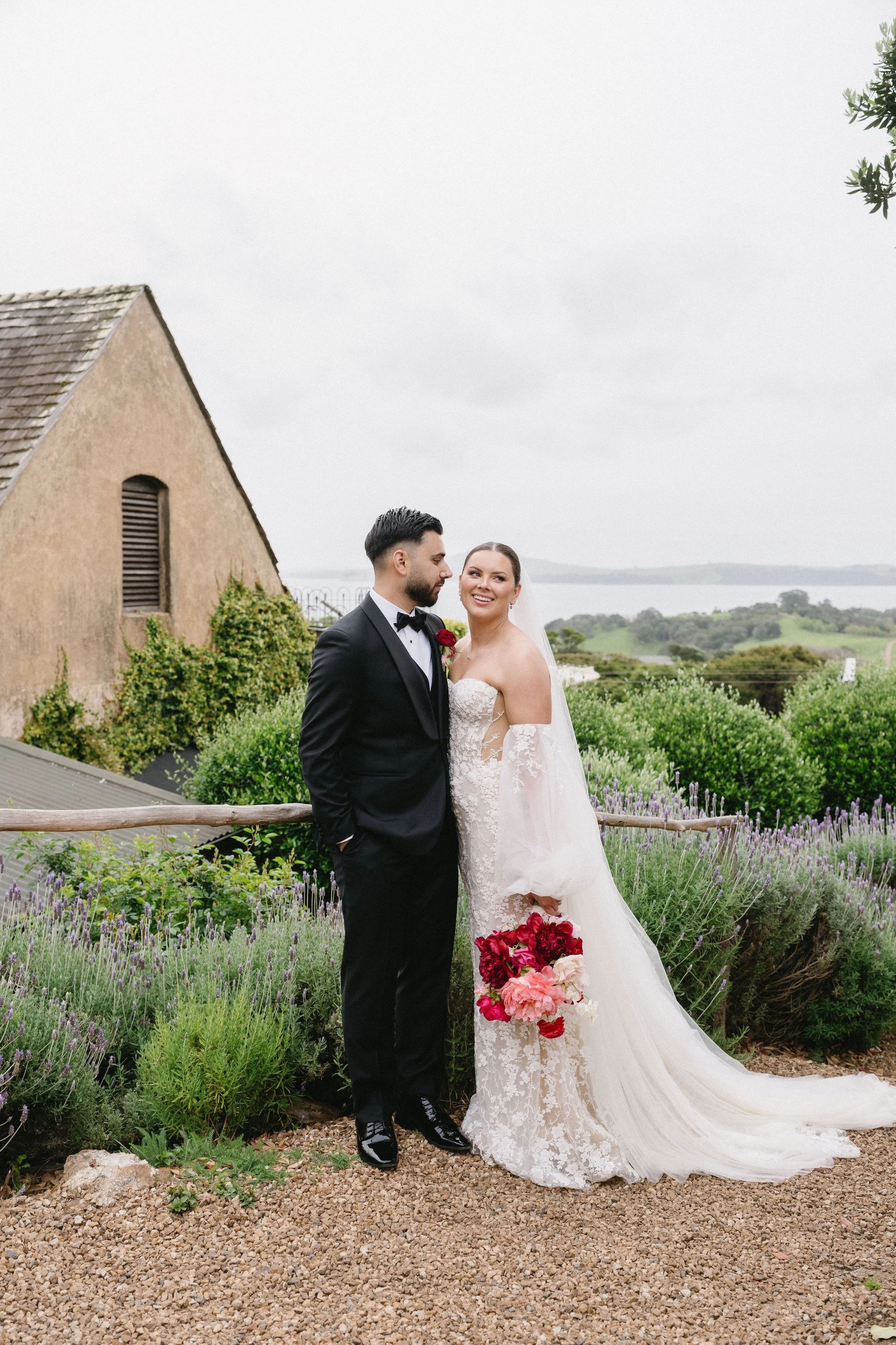 A bride and groom standing outdoors, with the bride holding a bouquet of red and pink flowers, on a gravel path surrounded by lavender and greenery, with a rustic building and a scenic landscape in the background.