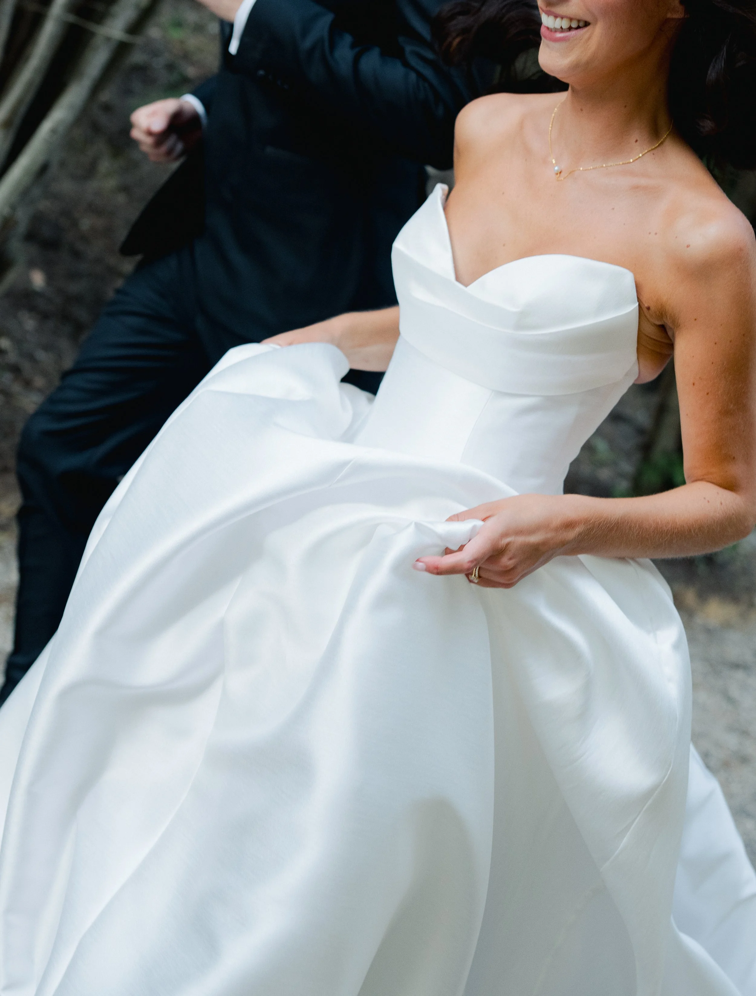 A woman in a white wedding dress smiling and holding the fabric of her dress, with a man in a black suit visible in the background.