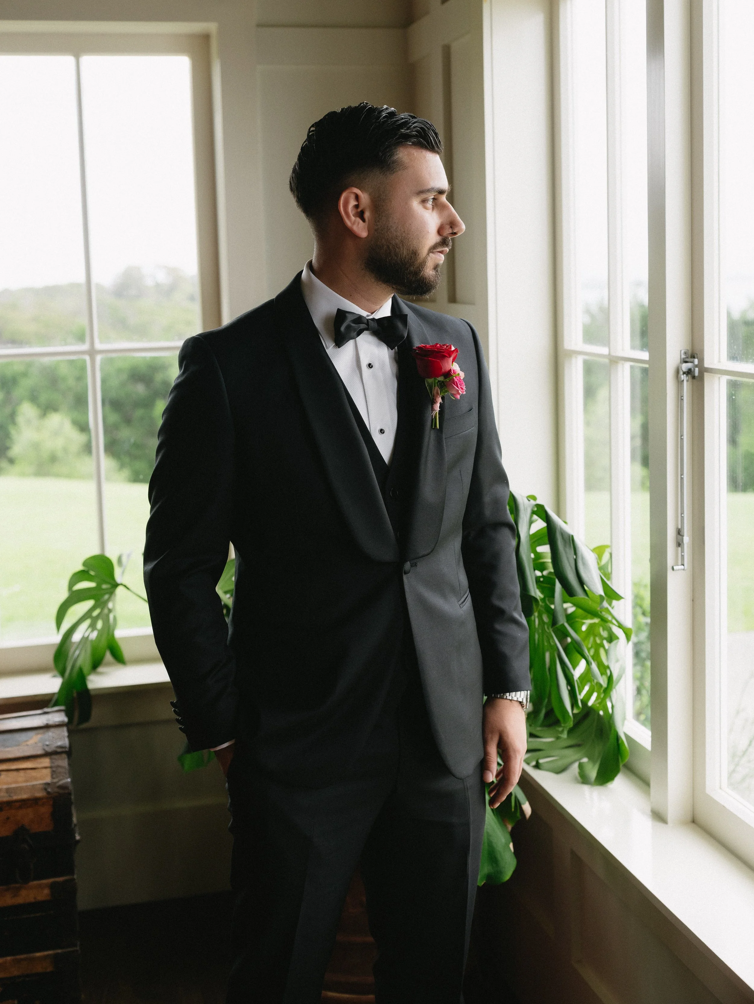 A groom in a black tuxedo with a bow tie and a red rose boutonniere, standing by a window and looking outside.