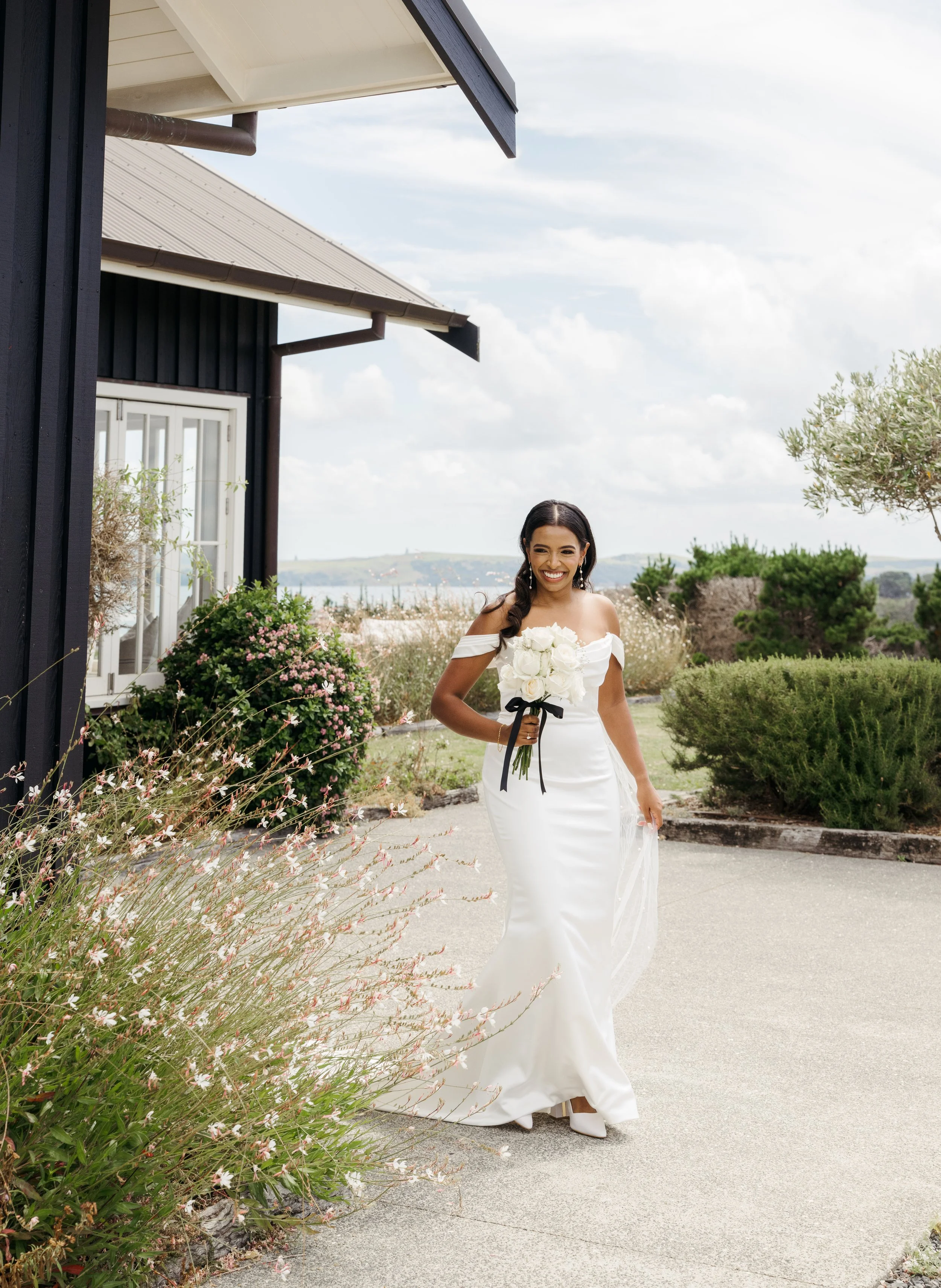 A woman in a white wedding dress holding a bouquet of white roses, walking outdoors near a black building with a garden and cloudy sky in the background.