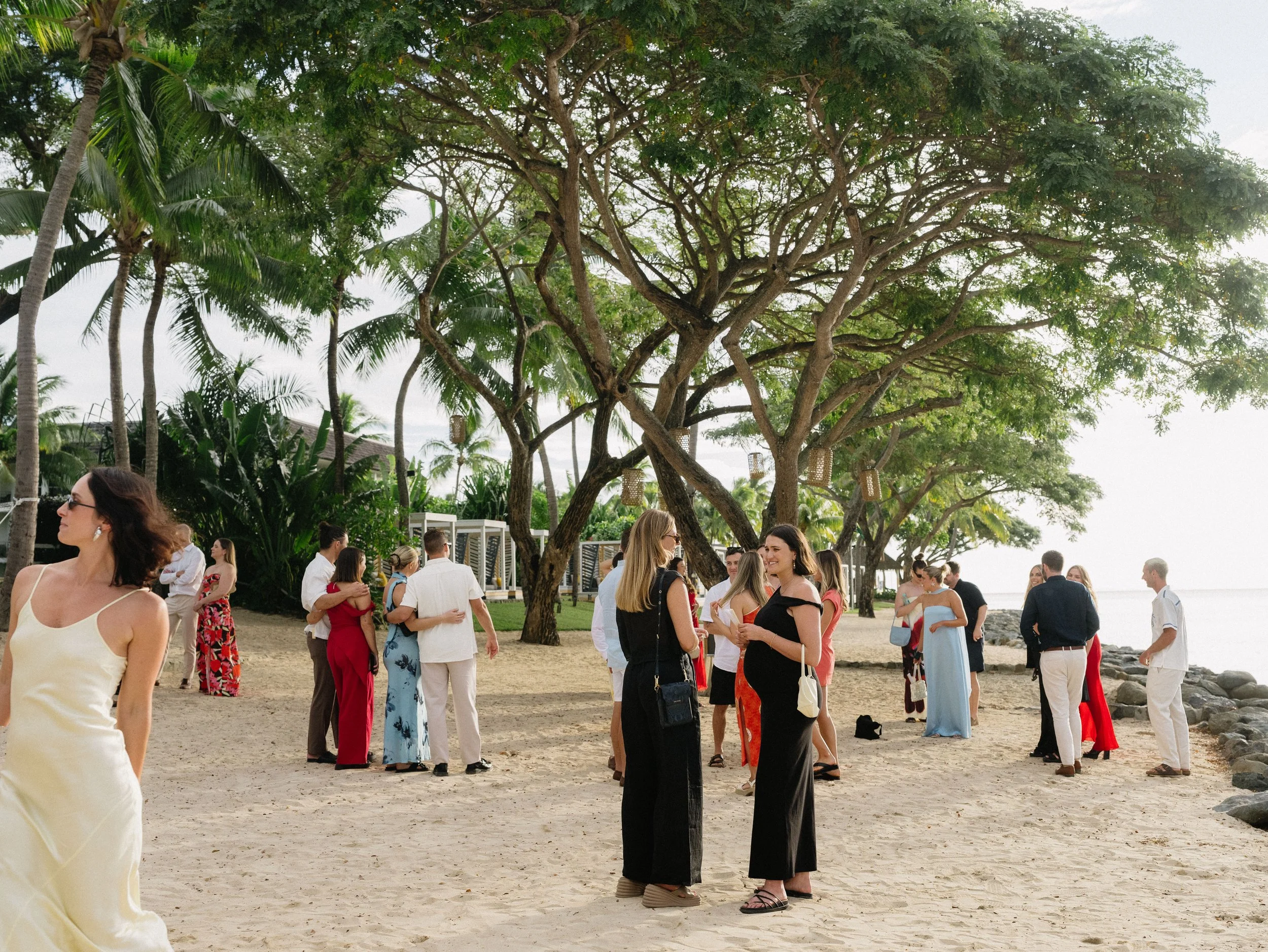 Group of people gathered on a sandy beach near large trees, with some dressed casually and others in formal attire, likely for a celebration or wedding.