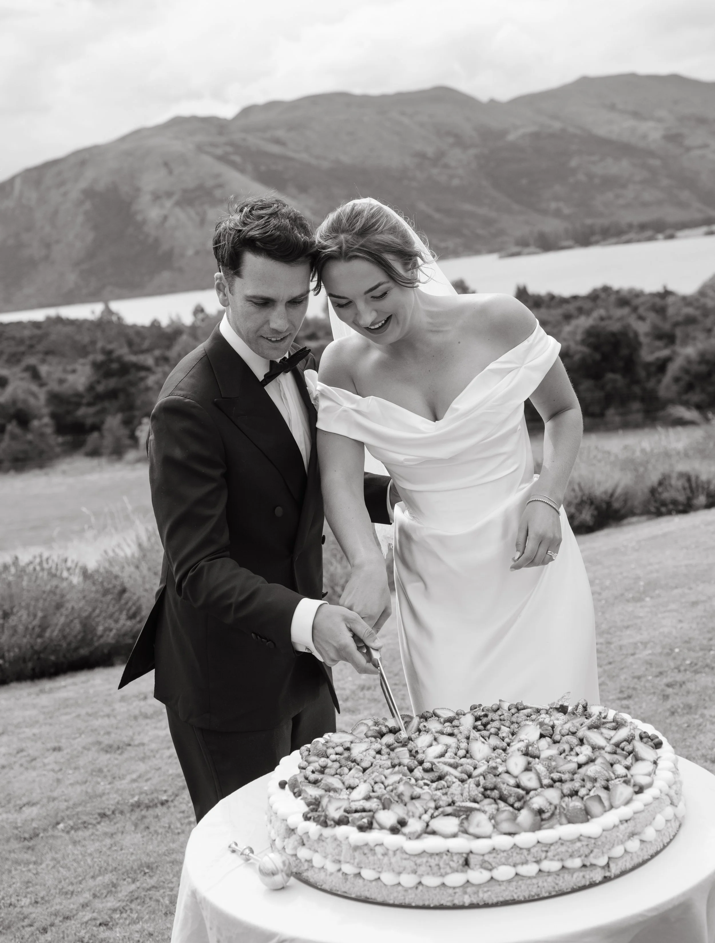 Black and white photo of a bride and groom cutting a large heart-shaped wedding cake outdoors with mountains in the background.