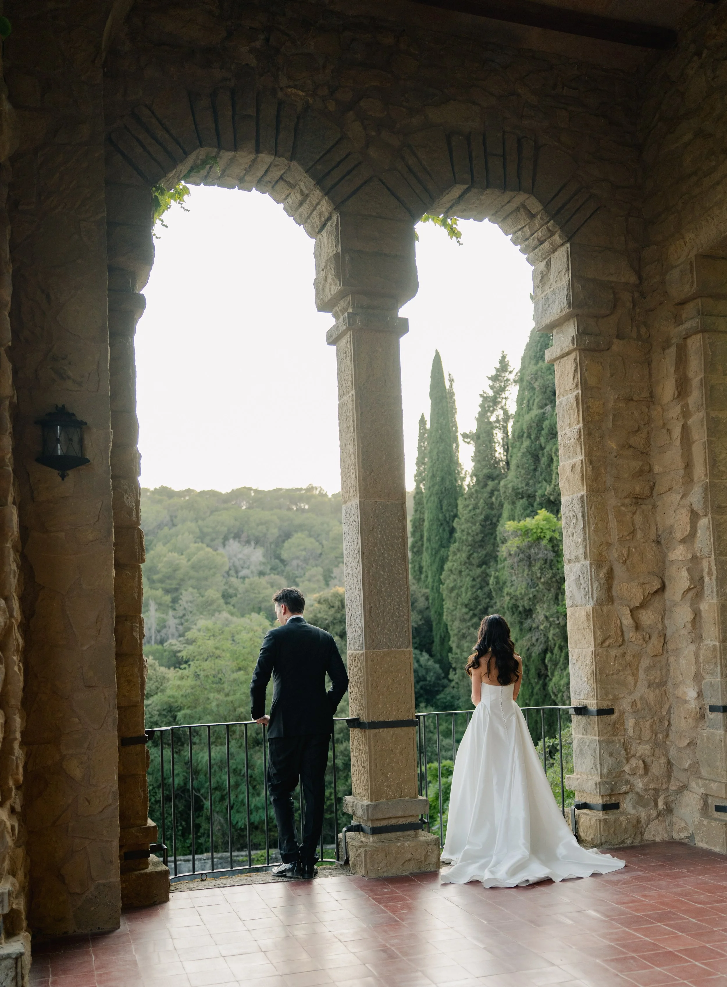 A man and a woman in wedding attire standing on a balcony framed by stone arches, overlooking a lush green landscape with trees.