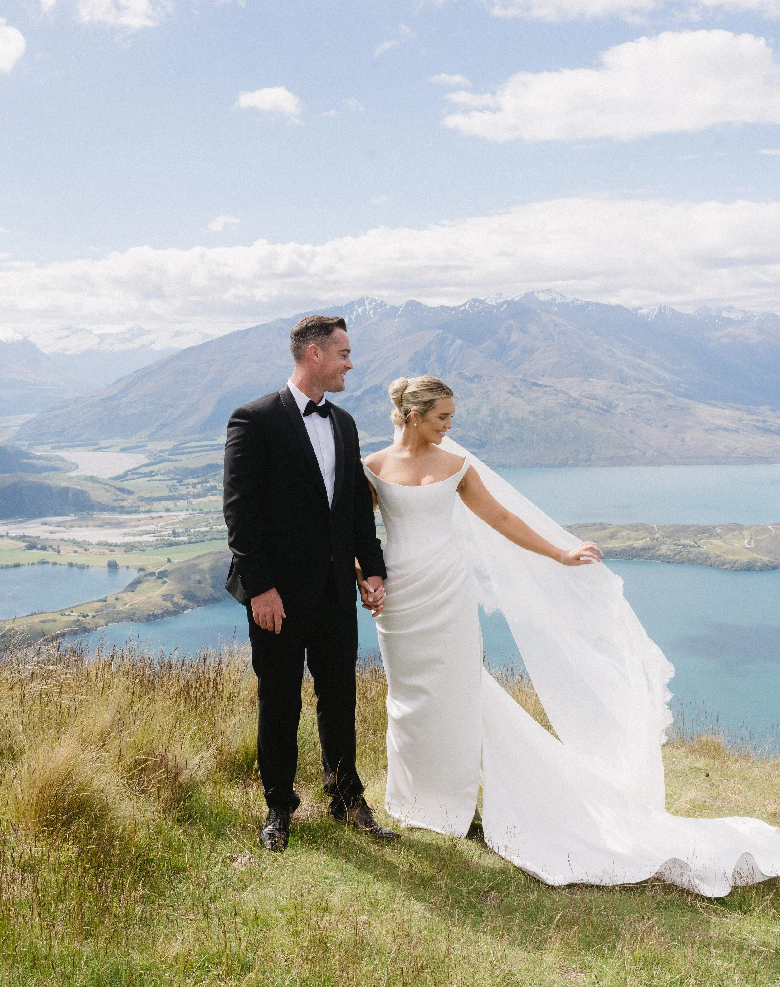 A bride and groom holding hands on a grassy hill with mountains, lakes, and clouds in the background.