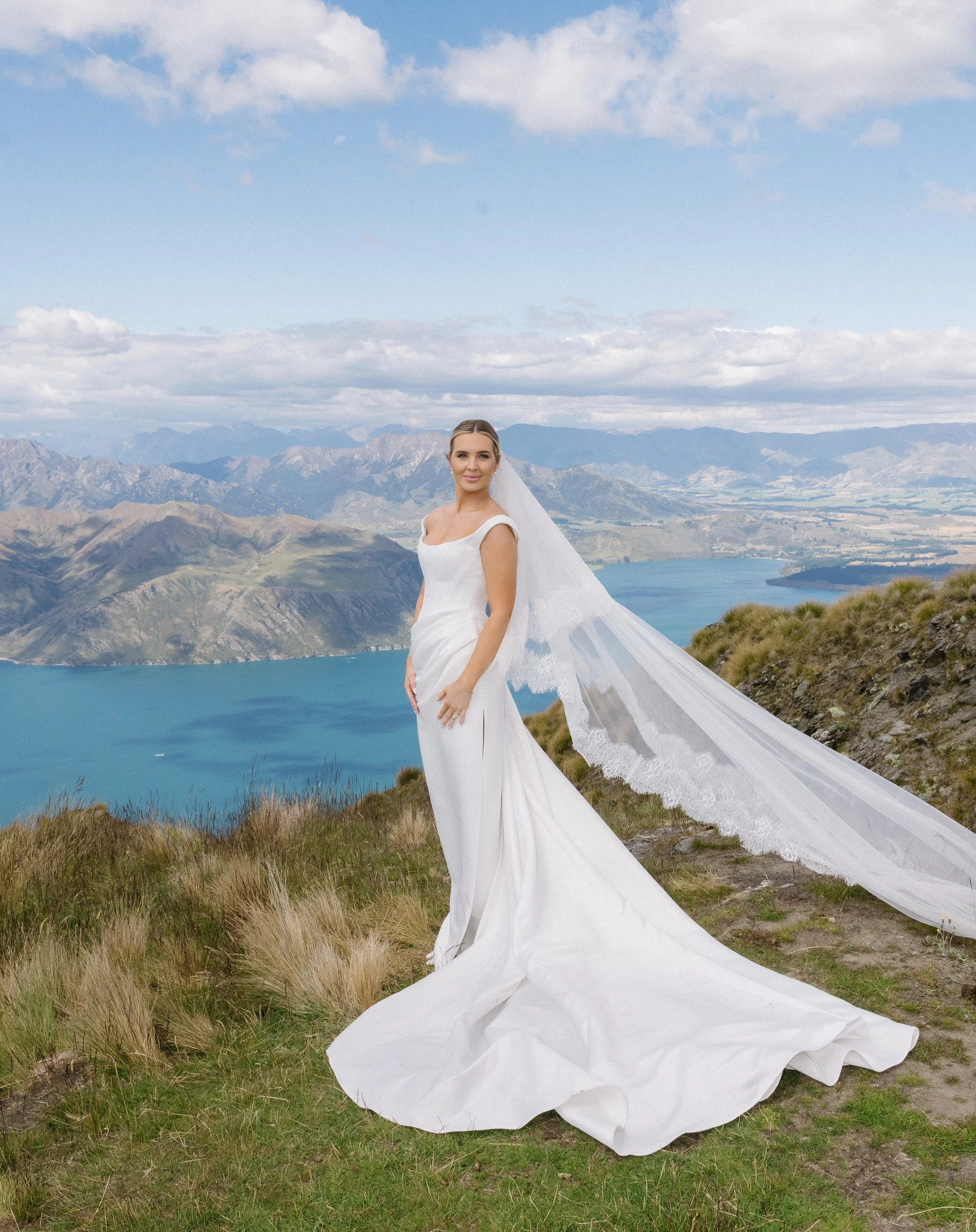 A bride standing outdoors on a grassy hill with mountains, a lake, and cloudy sky in the background, wearing a white wedding gown and veil.