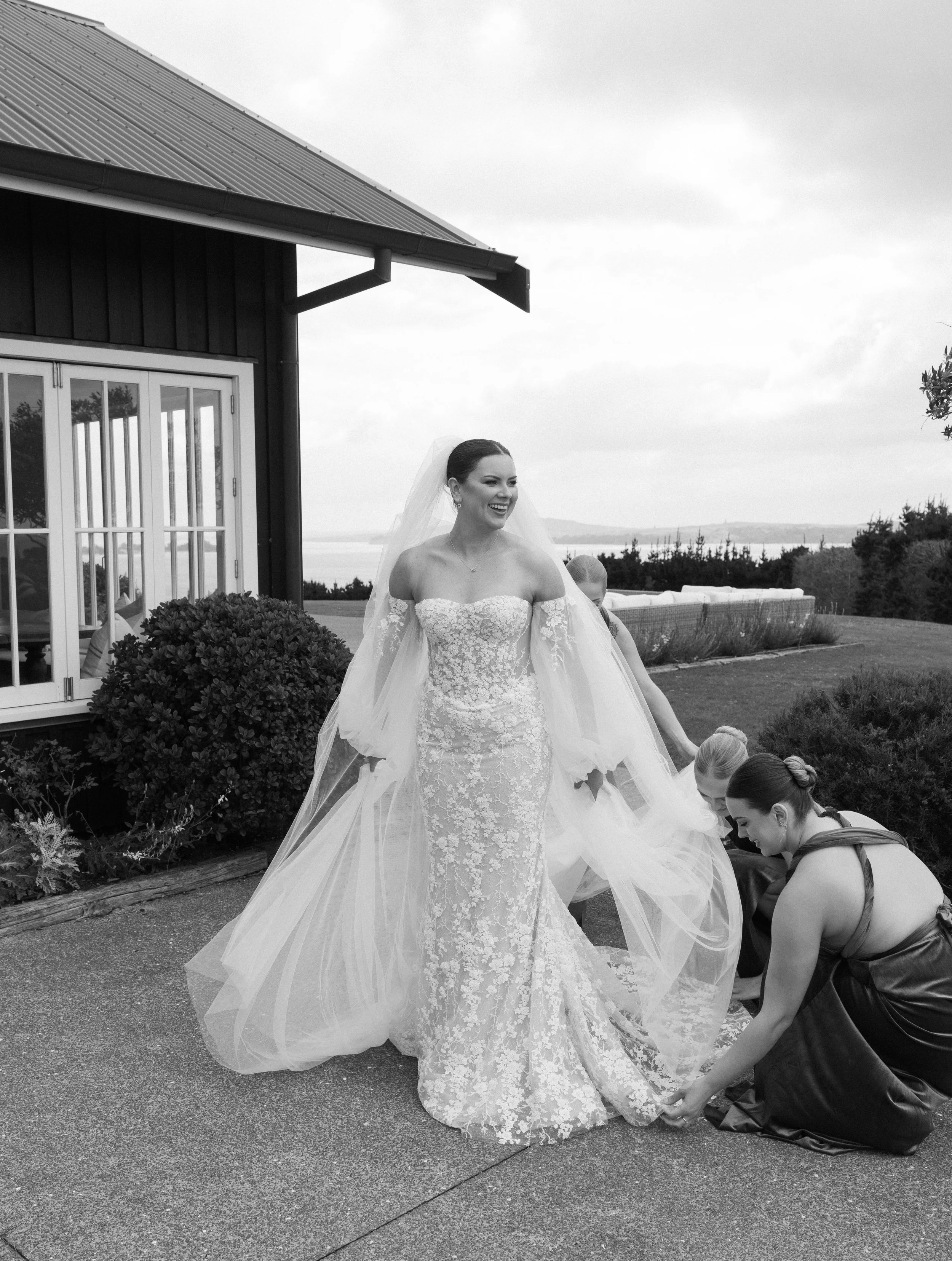 Black and white photo of a bride in a lace wedding gown and veil, smiling as she is helped with her dress outside a house, with a garden and scenic landscape in the background.