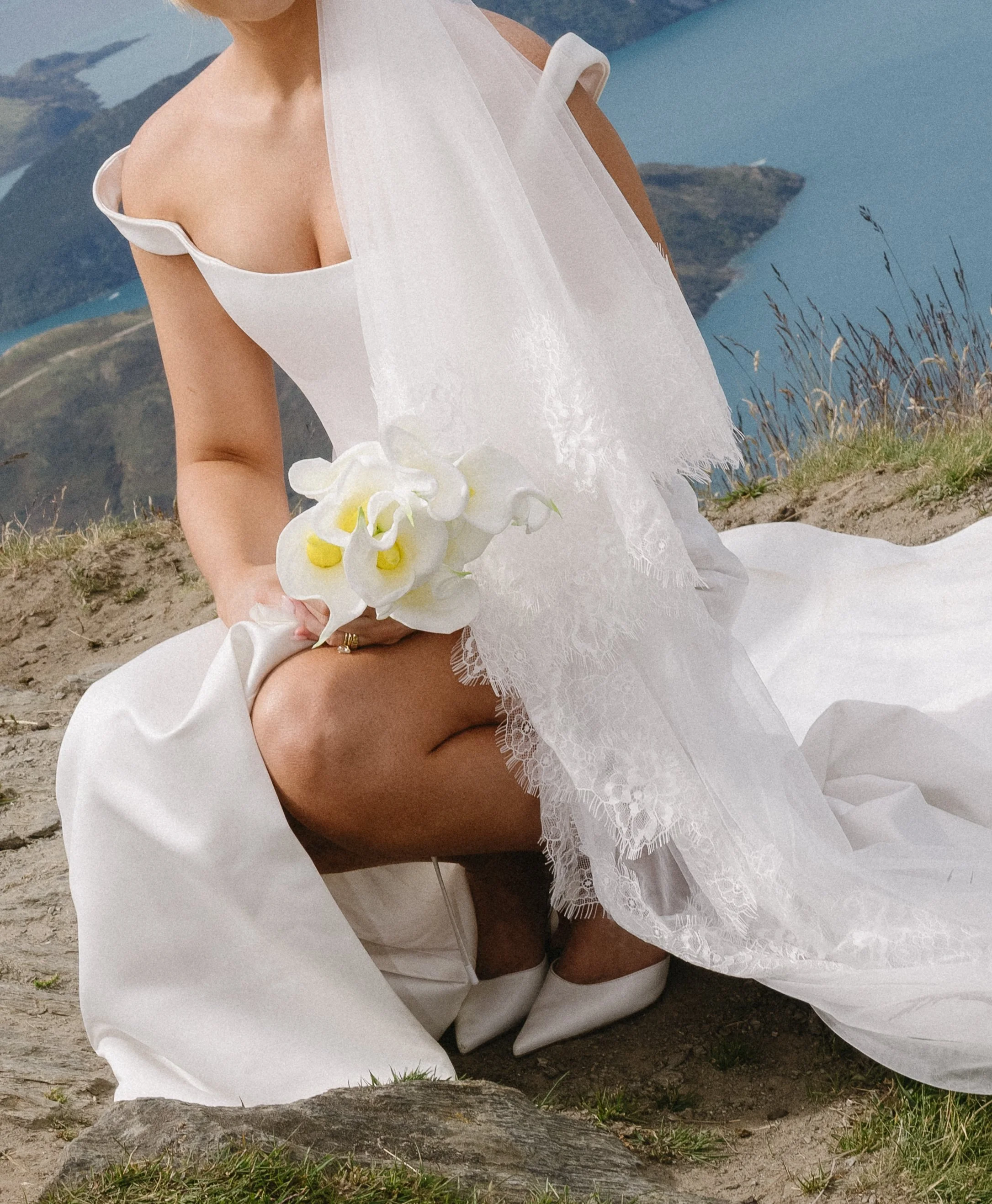 A woman dressed in a white wedding gown sitting on a rocky hilltop, holding a small bouquet of white flowers with yellow centers, with a scenic landscape of water and hills in the background.
