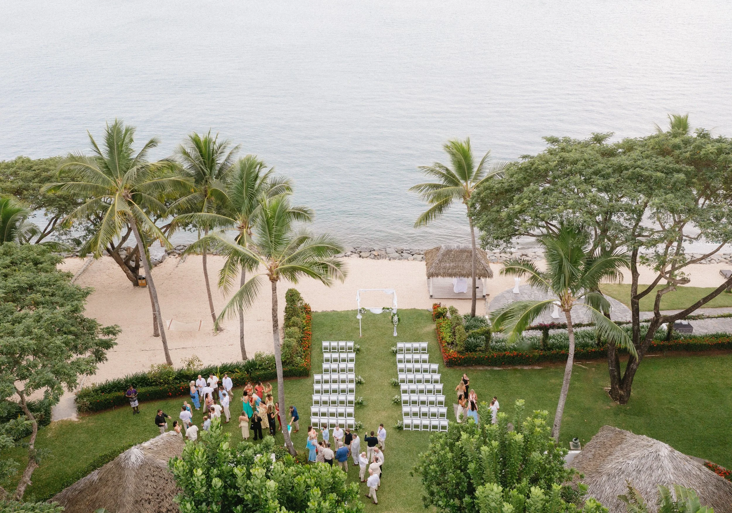 Beachside wedding setup with white chairs arranged in rows, an arch decorated with flowers, and guests gathering, surrounded by palm trees and lush greenery.