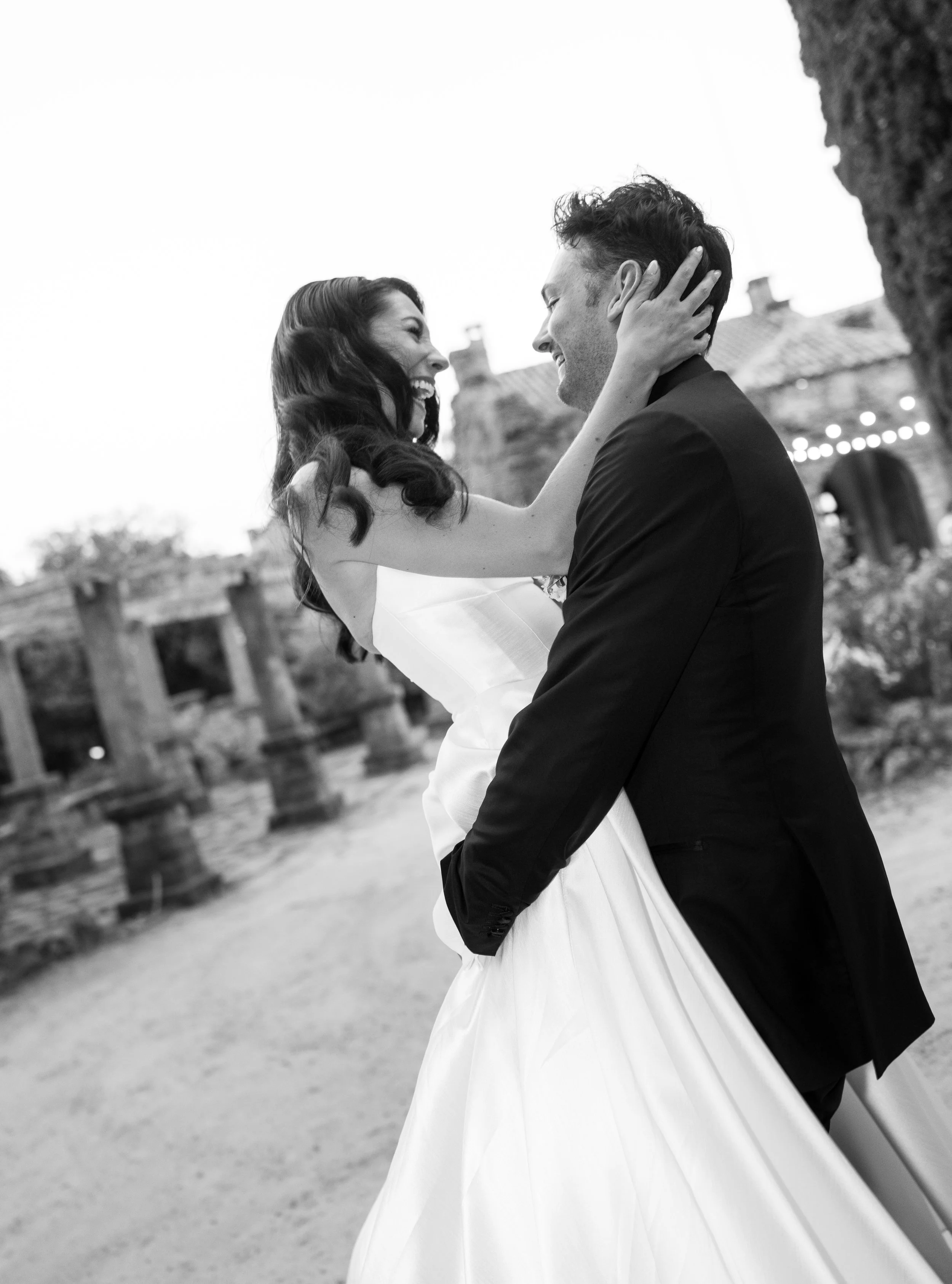 A black-and-white photo of a joyful bride and groom on their wedding day, with the bride lifting the groom and both smiling lovingly.