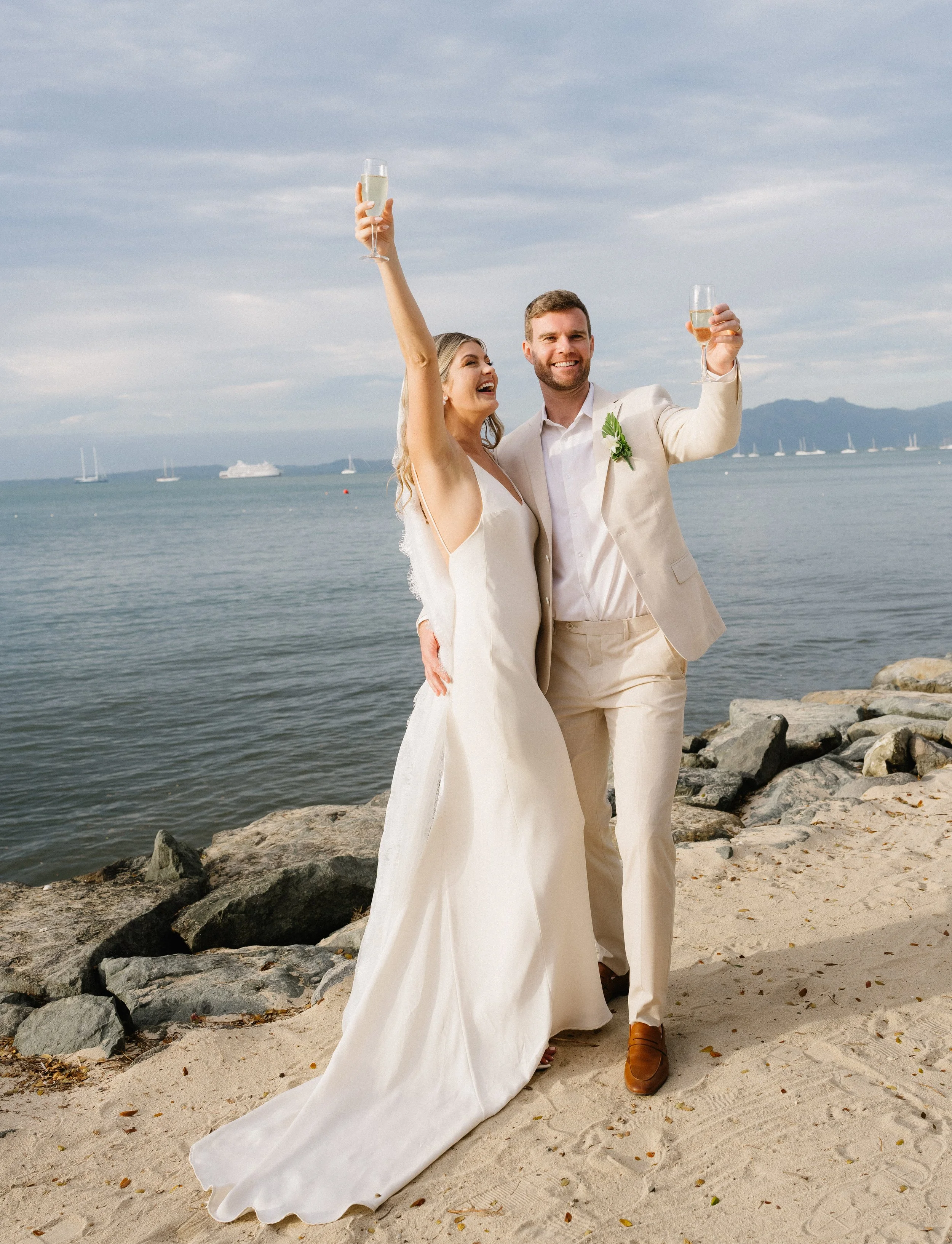 A newlywed couple celebrating with champagne on a beach, with boats in the water and mountains in the background.