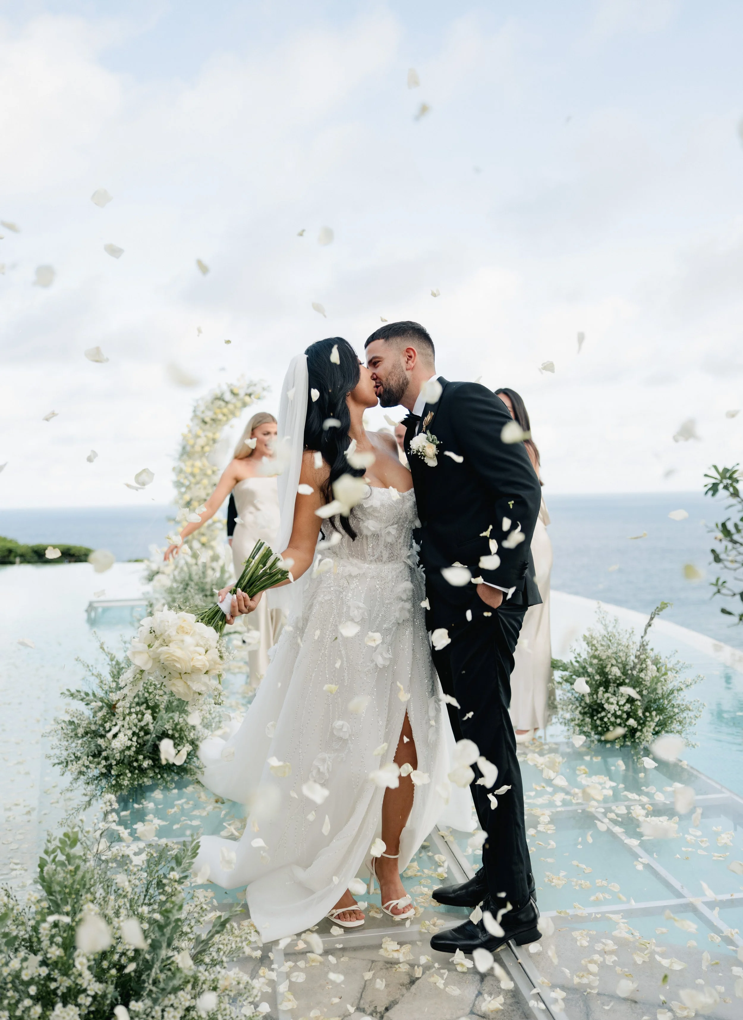 A bride and groom kiss at their wedding ceremony on a scenic outdoor location overlooking the ocean, surrounded by flower arrangements and falling white flower petals.