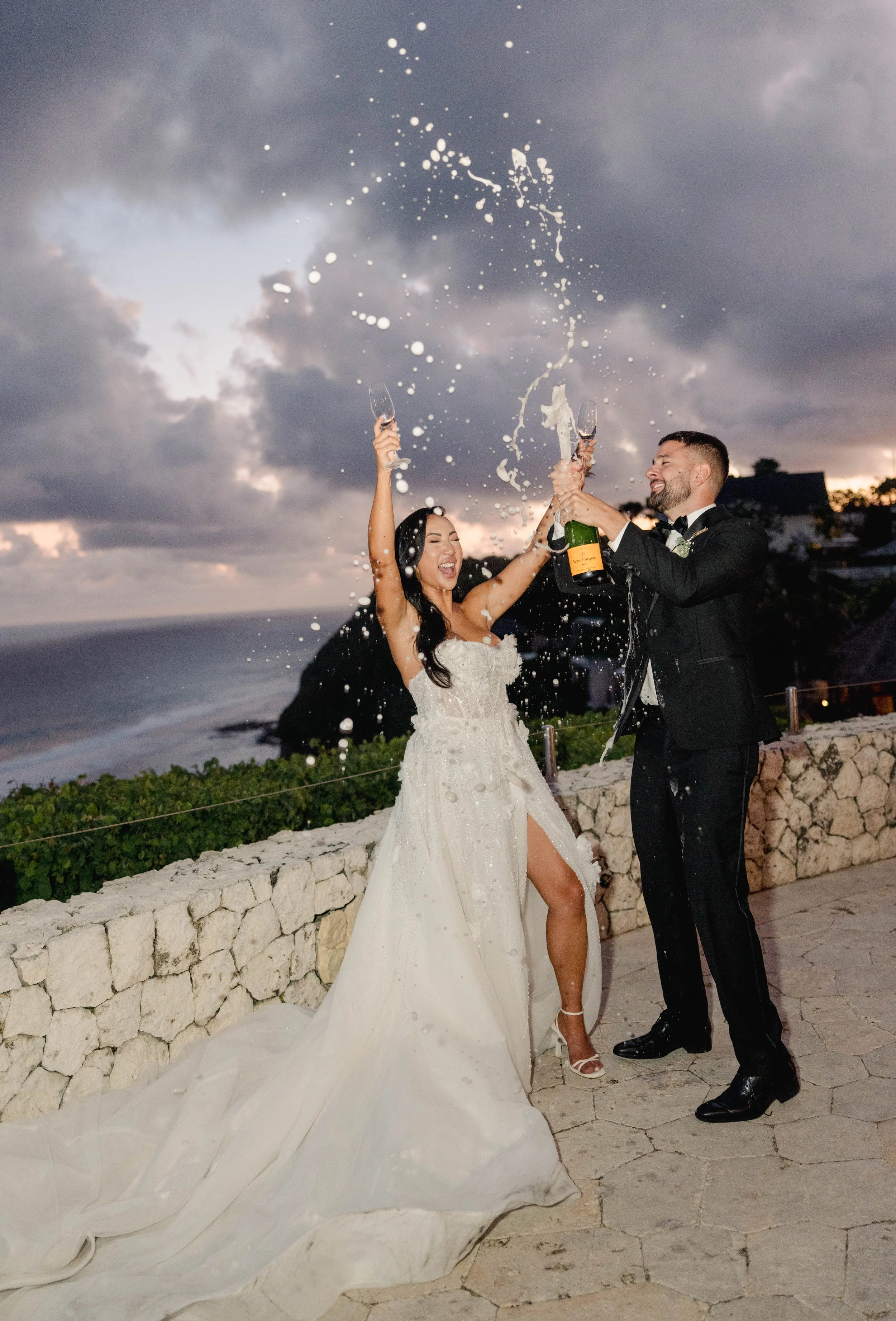 A newlywed couple celebrating with champagne on a terrace overlooking the ocean at sunset. The bride is wearing a white wedding dress and the groom is in a black tuxedo. Both are smiling and holding champagne glasses amidst a splash of champagne.