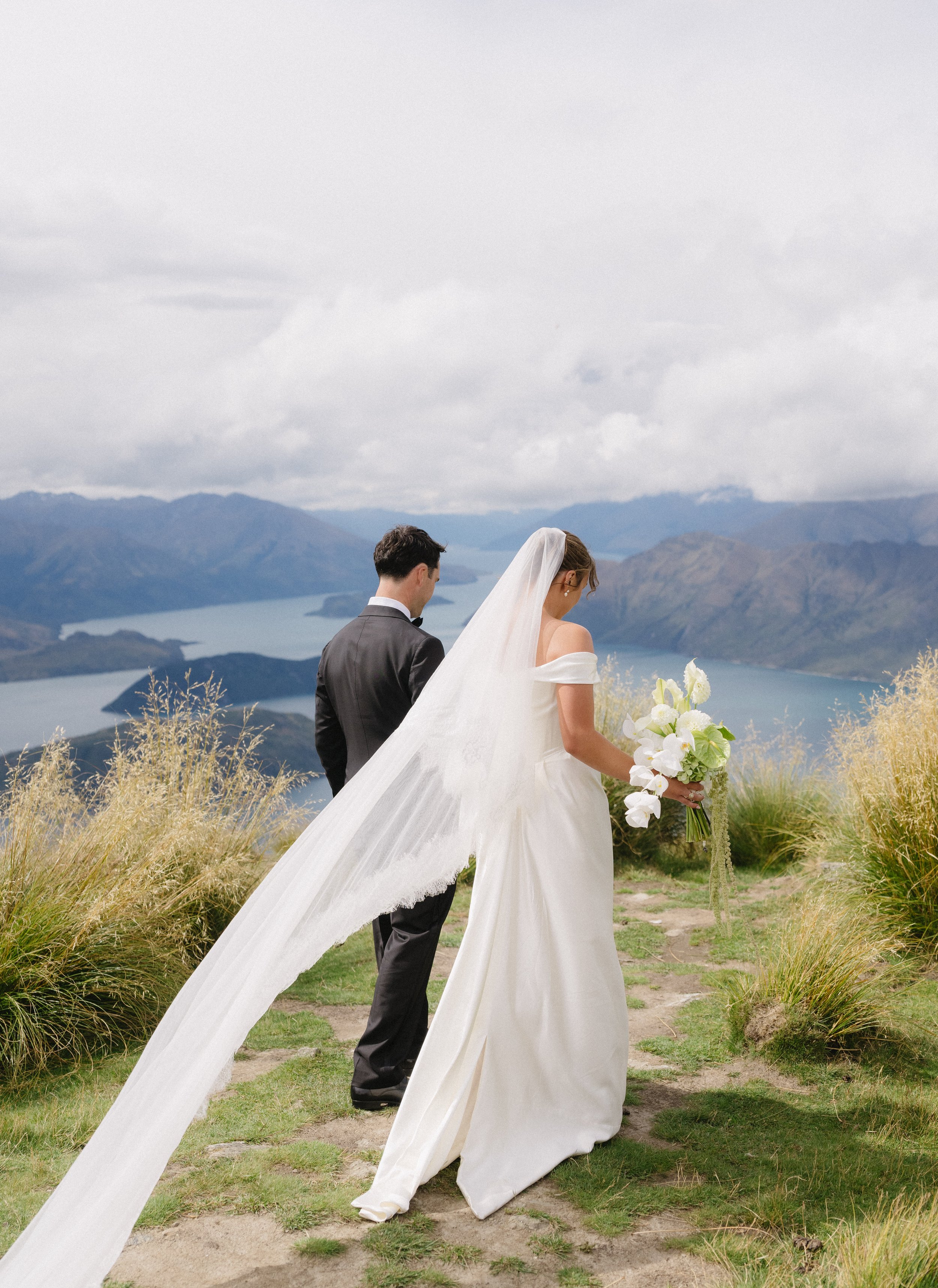 Bride in a white wedding dress and veil holding a bouquet, standing on a grassy trail with a groom in a black suit, overlooking a mountain lake landscape under cloudy sky.